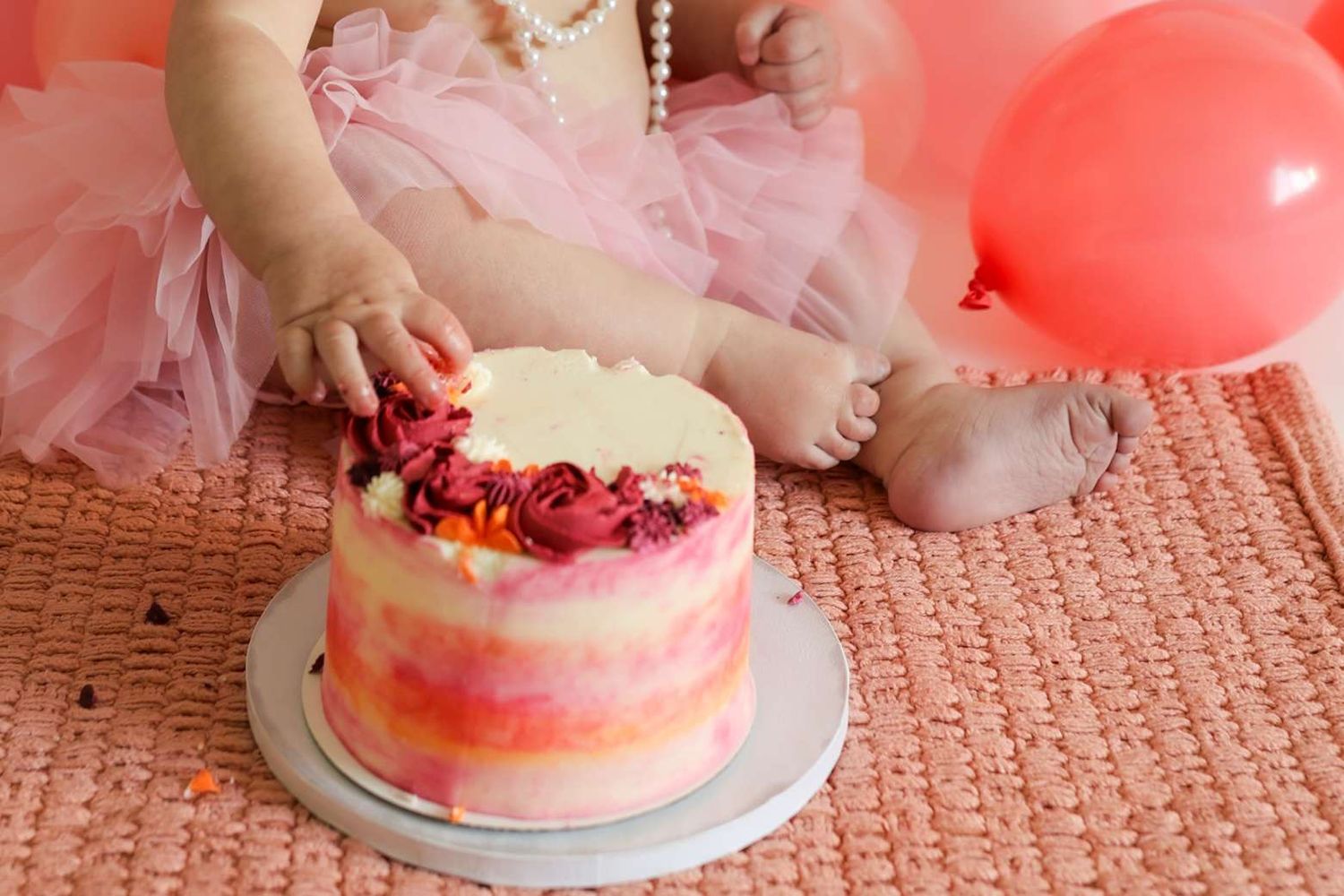 A pink and white smash cake being destroyed by small hands on a pink carpet during a first birthday celebration.