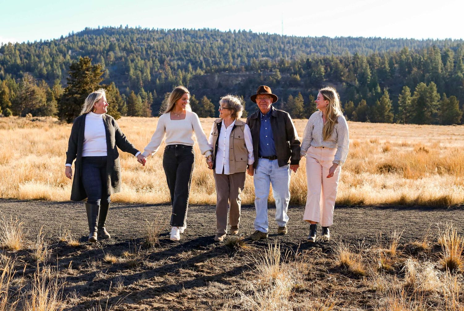 A group walks together through a golden field with evergreen trees in the background at sunset.