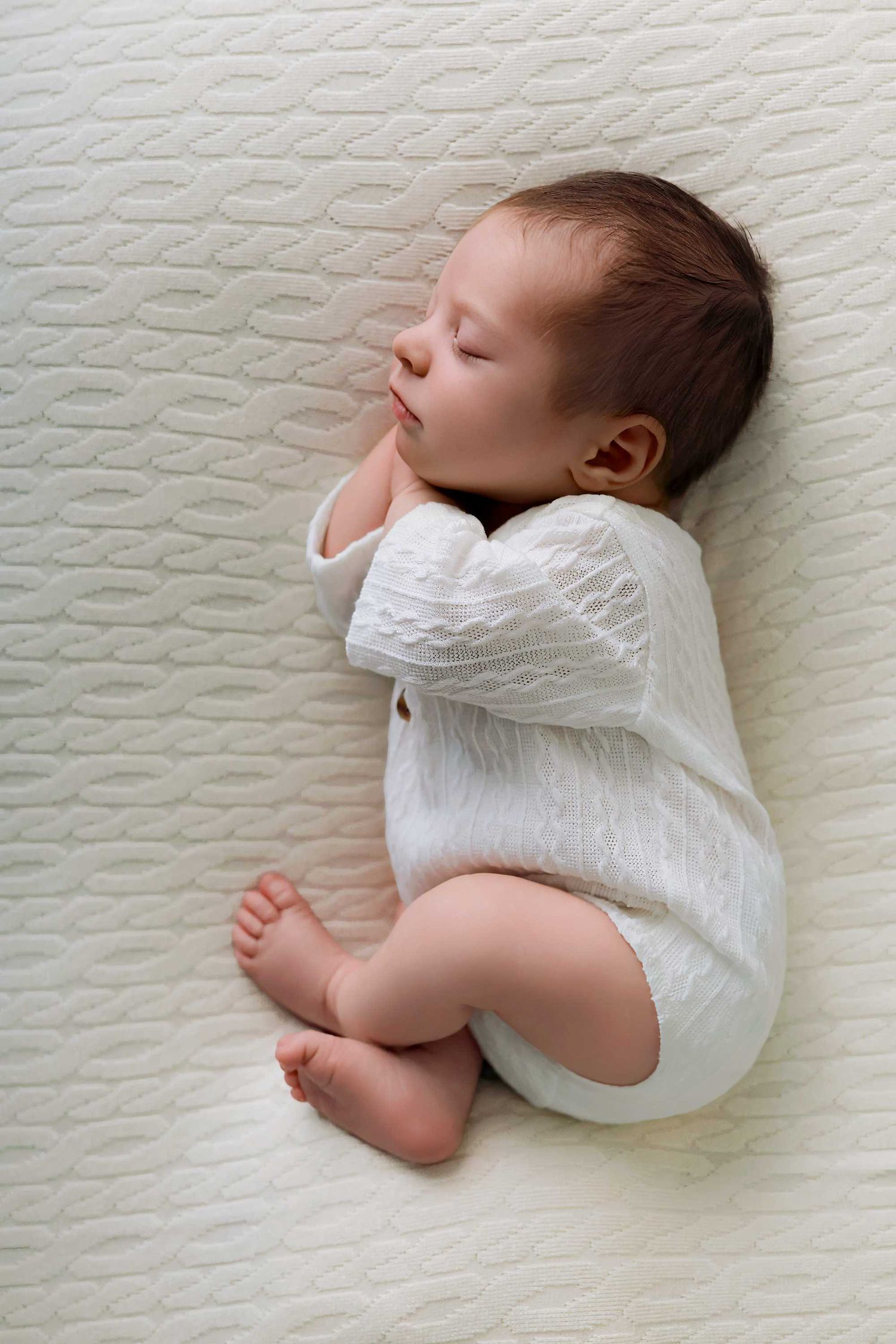Sleeping infant in white onesie lying on textured white blanket with tiny feet showing.