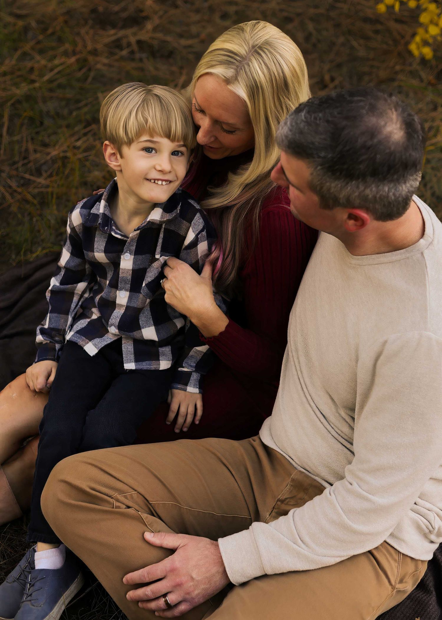 A young boy in a plaid shirt sits smiling with his blonde mother and father during an outdoor fall family portrait.