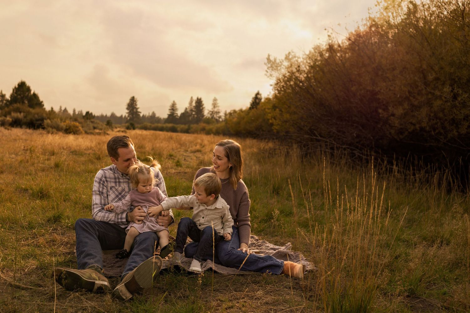 Family of four sitting together in a golden autumn field at sunset, sharing a joyful moment outdoors. Near Bend Oregon