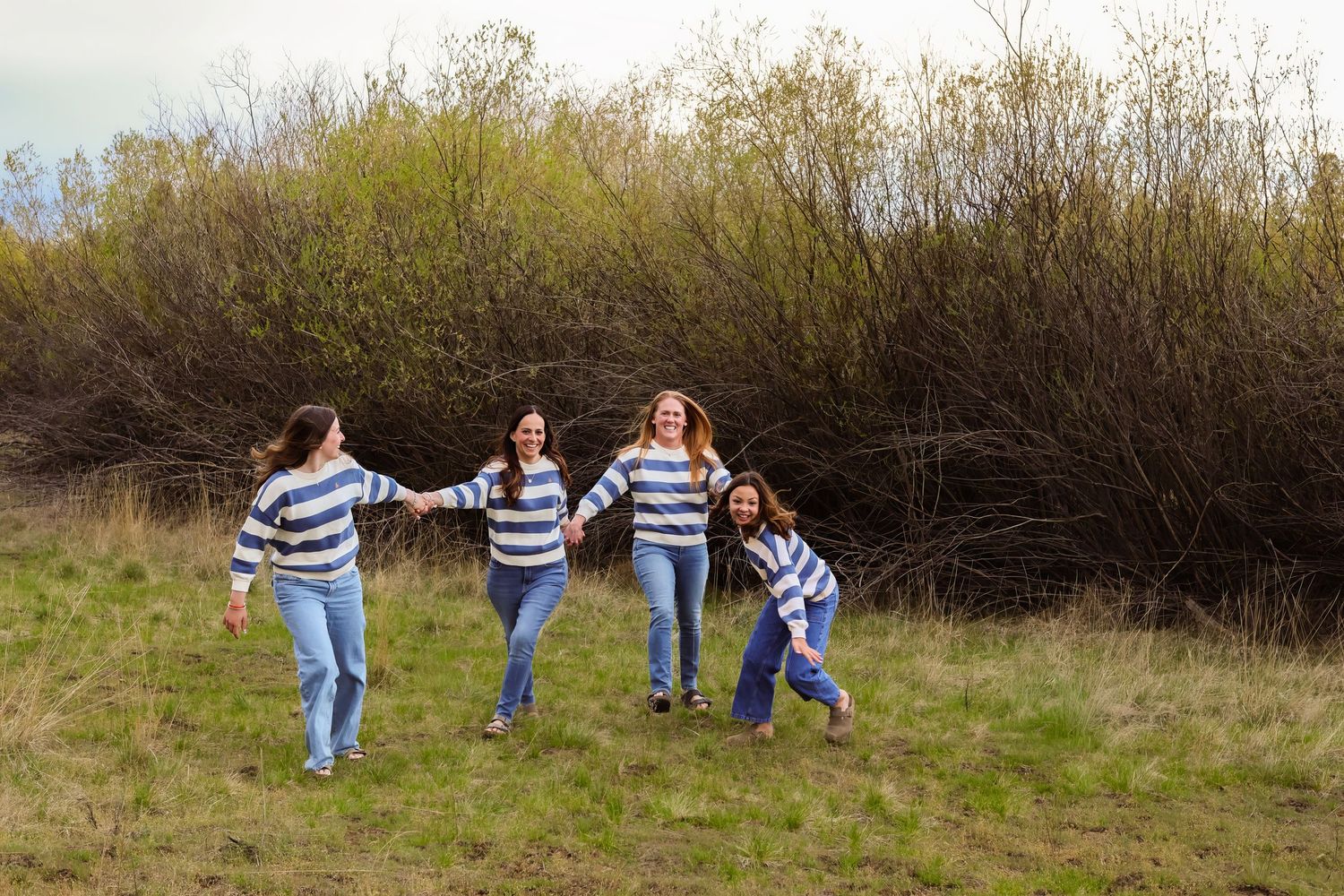 Four women in matching striped sweaters and jeans hold hands while running through a green meadow.
