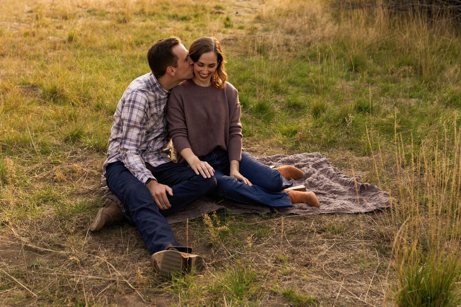 Couple sitting on blanket in golden field, man whispering to smiling woman, casual outdoor engagement photo.