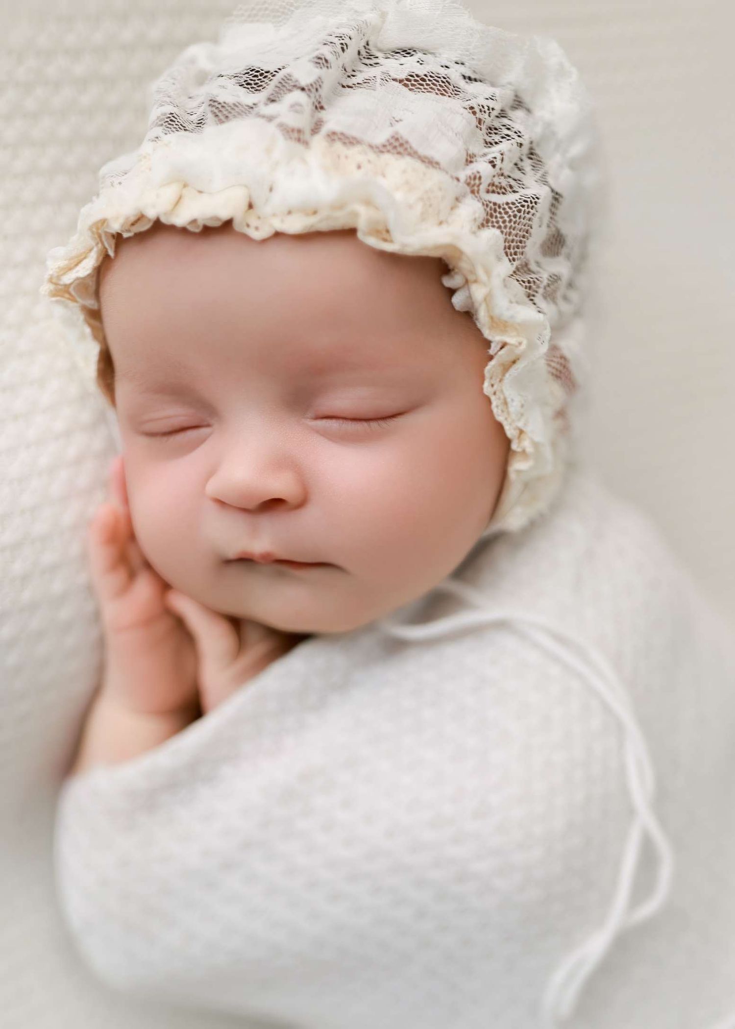 Peaceful newborn wearing delicate lace bonnet while sleeping on soft white fabric.