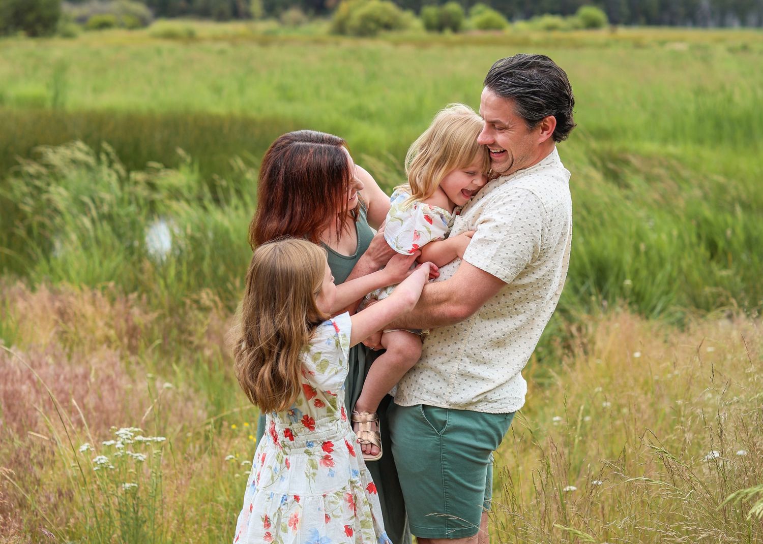 Family of four sharing a joyful embrace in a lush green meadow, parents holding young daughters outdoors.