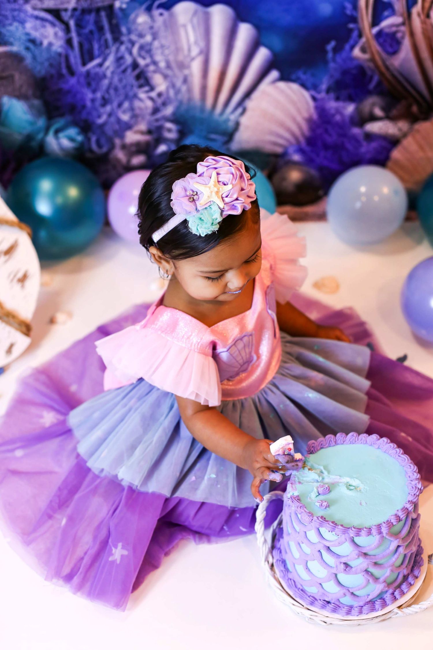 Toddler girl in purple pink tutu dress reaching for mermaid themed birthday cake with ocean decor.