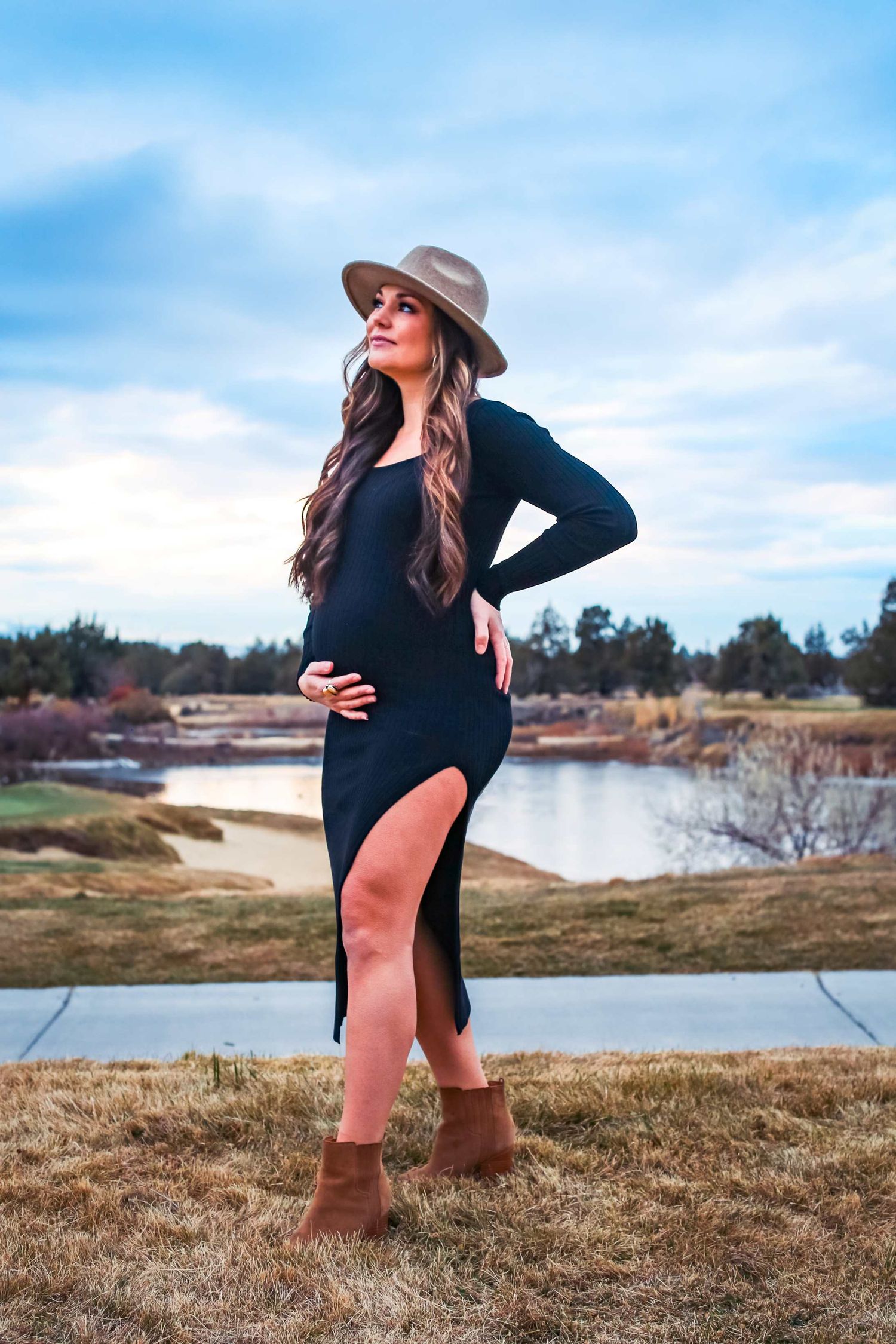 Pregnant woman in a black slit dress and tan hat posing outdoors near a lake.