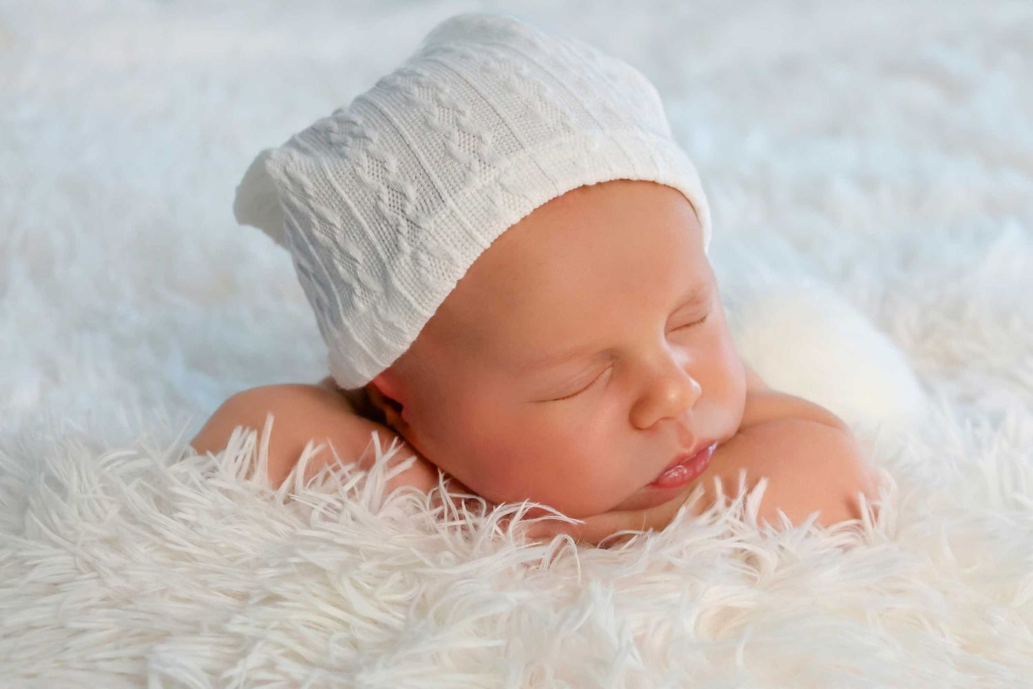 Peaceful sleeping newborn wearing white cap rests on soft white furry blanket.