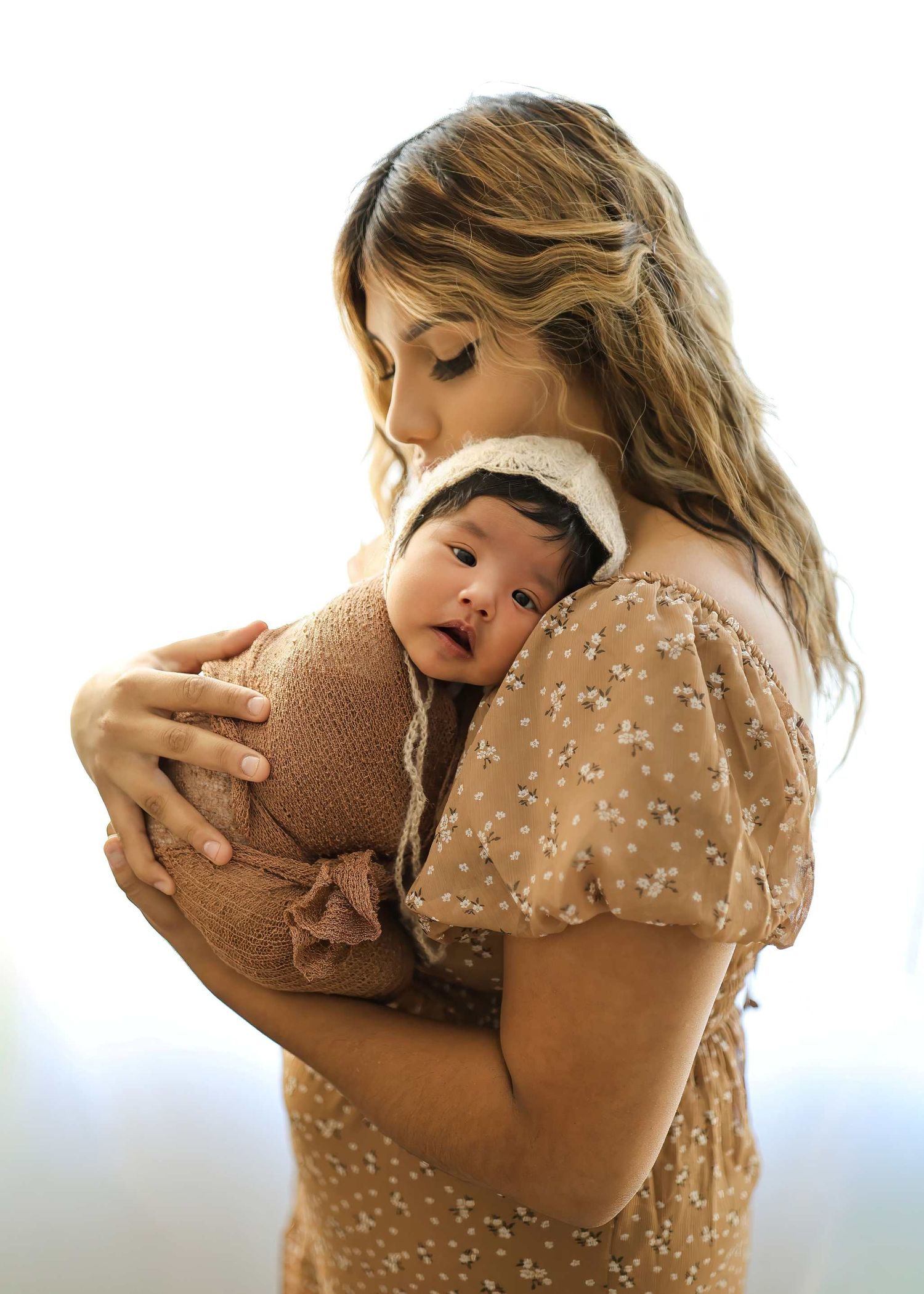 A mother in a floral dress tenderly holds her swaddled newborn baby wearing a white lace bonnet.