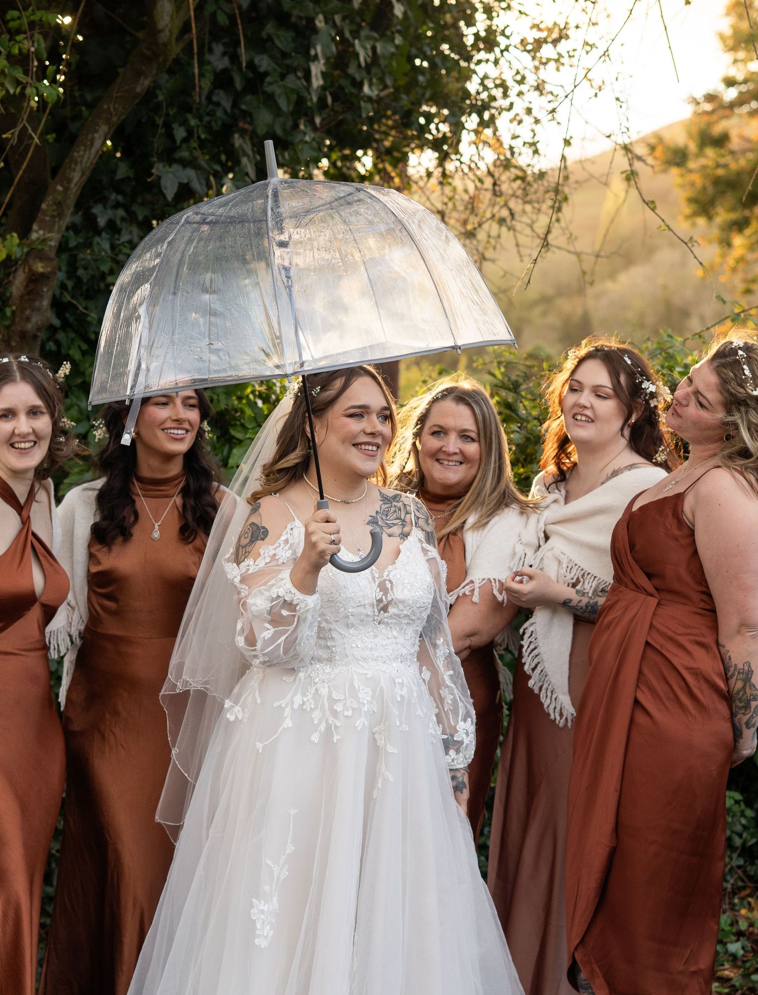 A bride with her bridesmaids, sharing an umbrella during golden hour rain.
