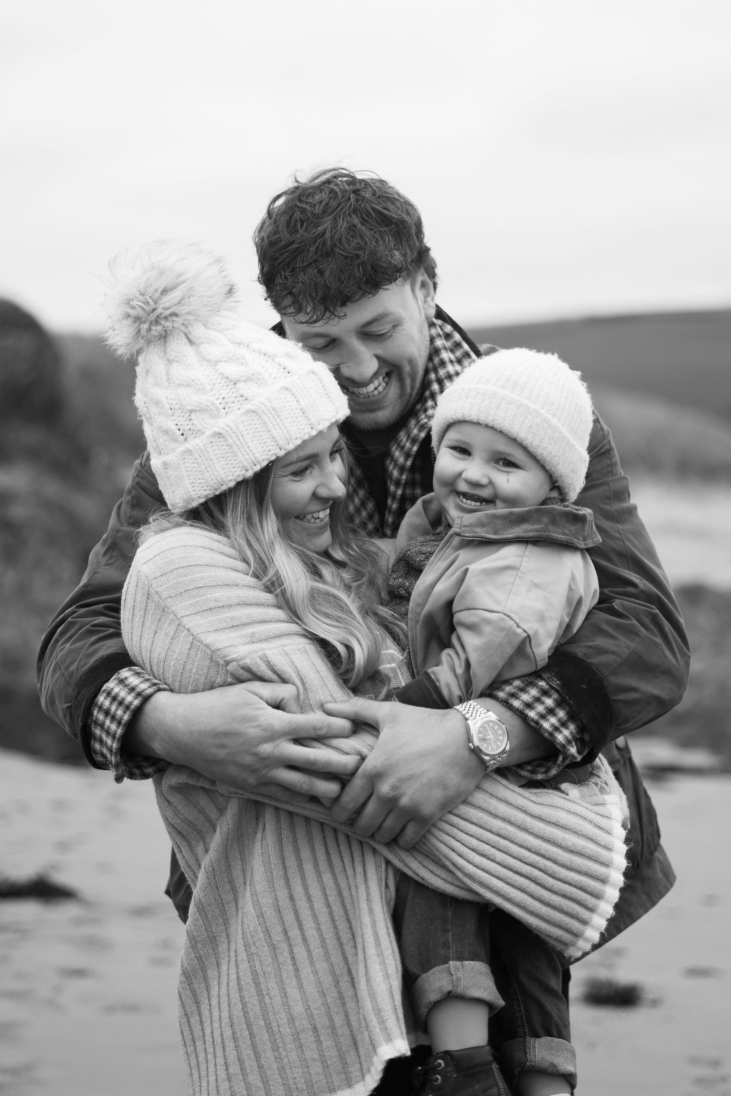 A mum, dad, and toddler son cuddle together on the beach during their family photoshoot on Bantham Beach, South Devon.