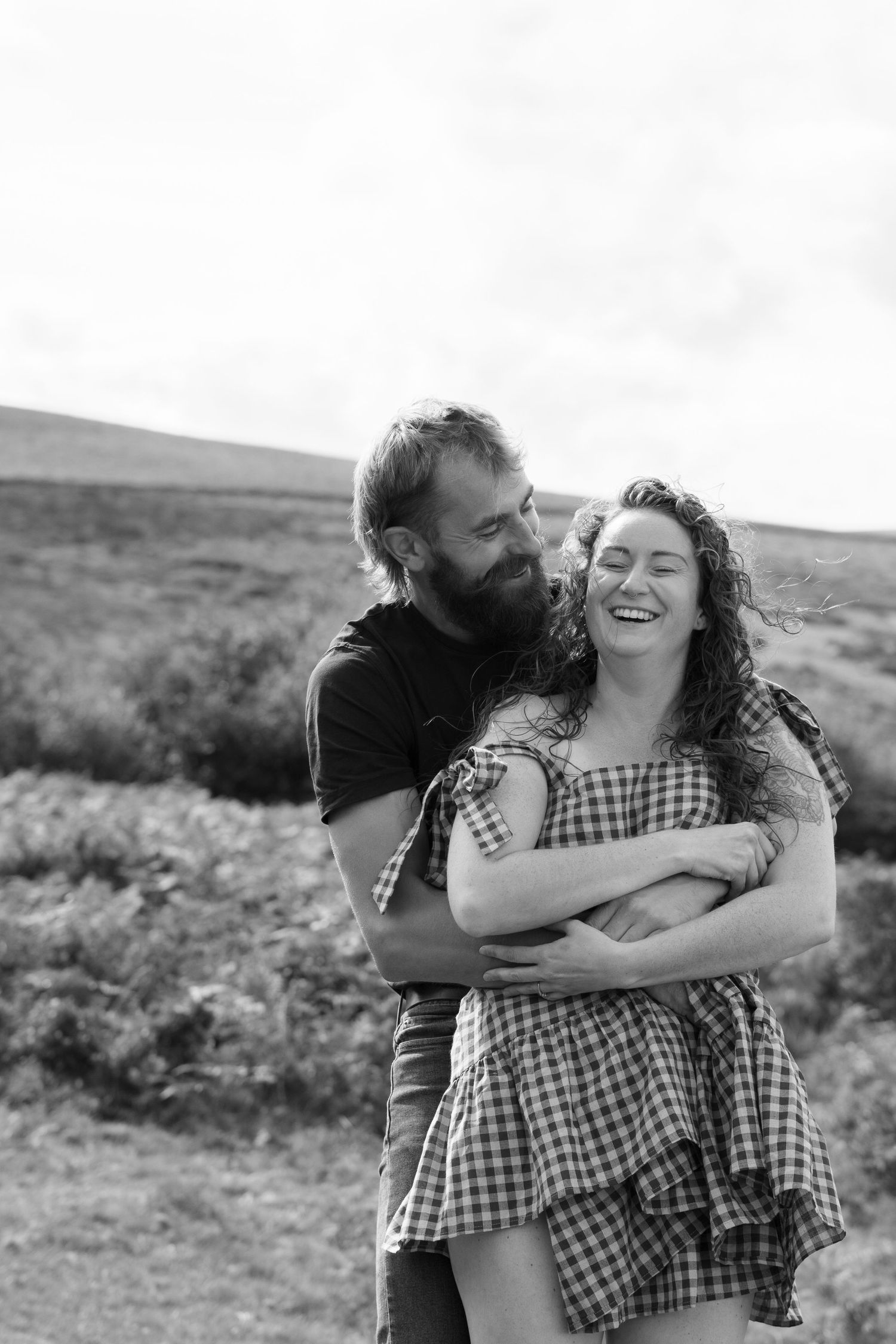 Black and white photo of a man cuddling his fiancé from behind at Dartmoor National Park, Devon, England, whilst both giggling.