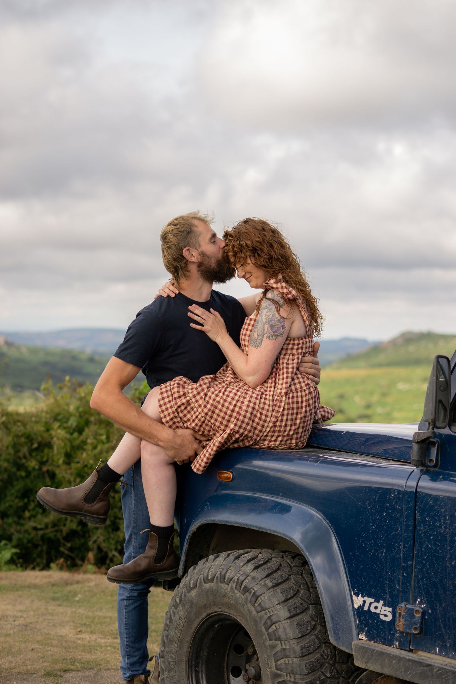A man kissing his fiancé on the forehead whilst she sits on the bonnet of their land rover at Dartmoor National Park, Devon, England.