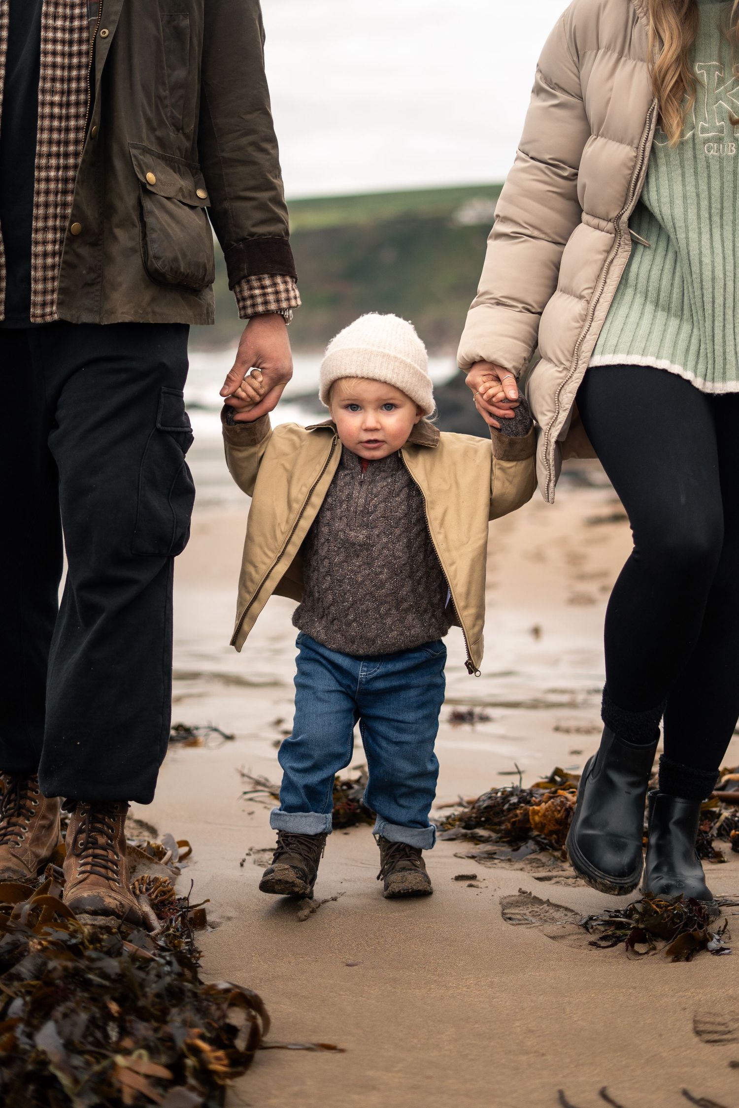 A mother and father hold hands with their toddler son during a family photo session on Bantham Beach, South Devon.