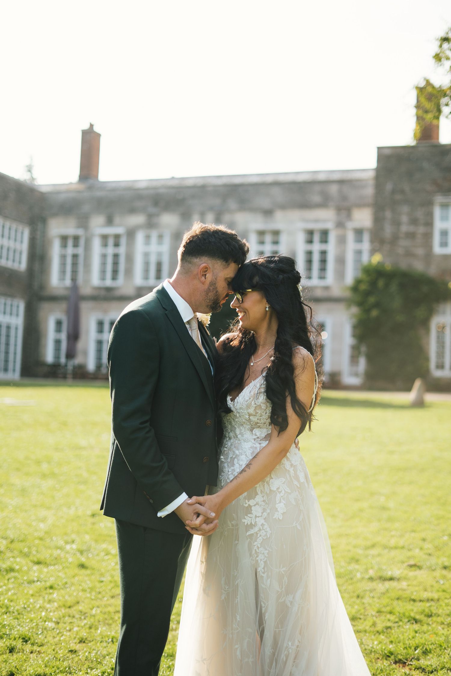 A bride and groom embracing with foreheads together, standing in front of their wedding venue (Cockington Court, Torquay, Devon).