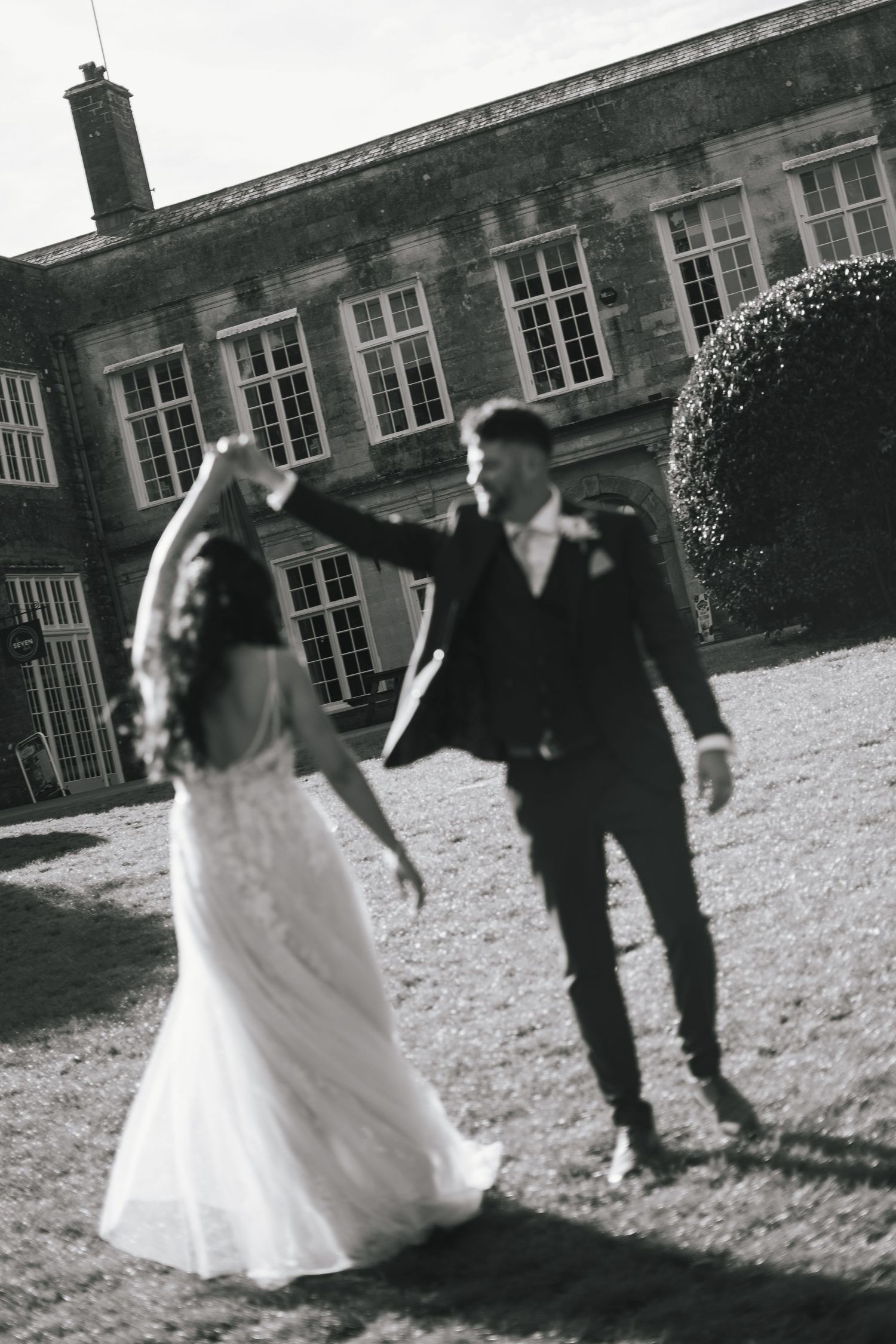 A black and white photo of a groom spinning his bride outside of Cockington Court, Torquay, Devon, England.