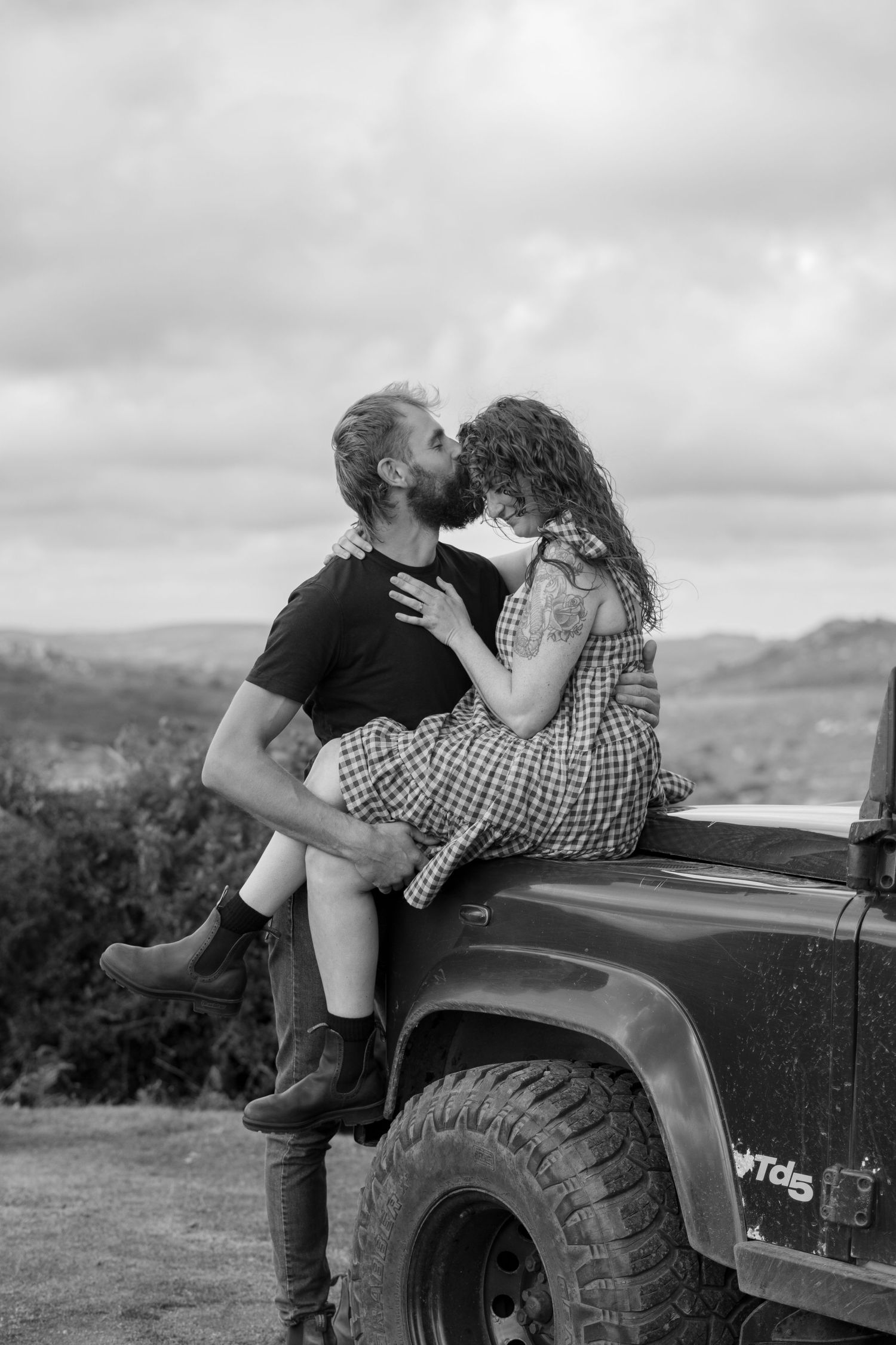Black and white photo of a man kissing his fiancé on the forehead whilst she sits on the bonnet of their land rover at Dartmoor, Devon