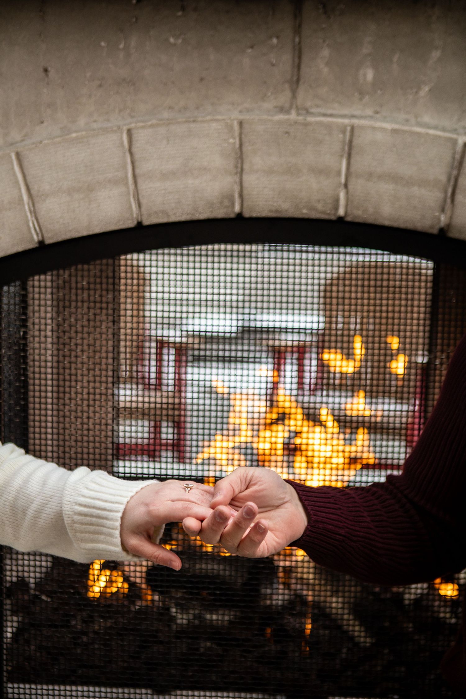 Hands warming up by a cozy fireplace seen through a mesh screen with glowing orange flames in the background.