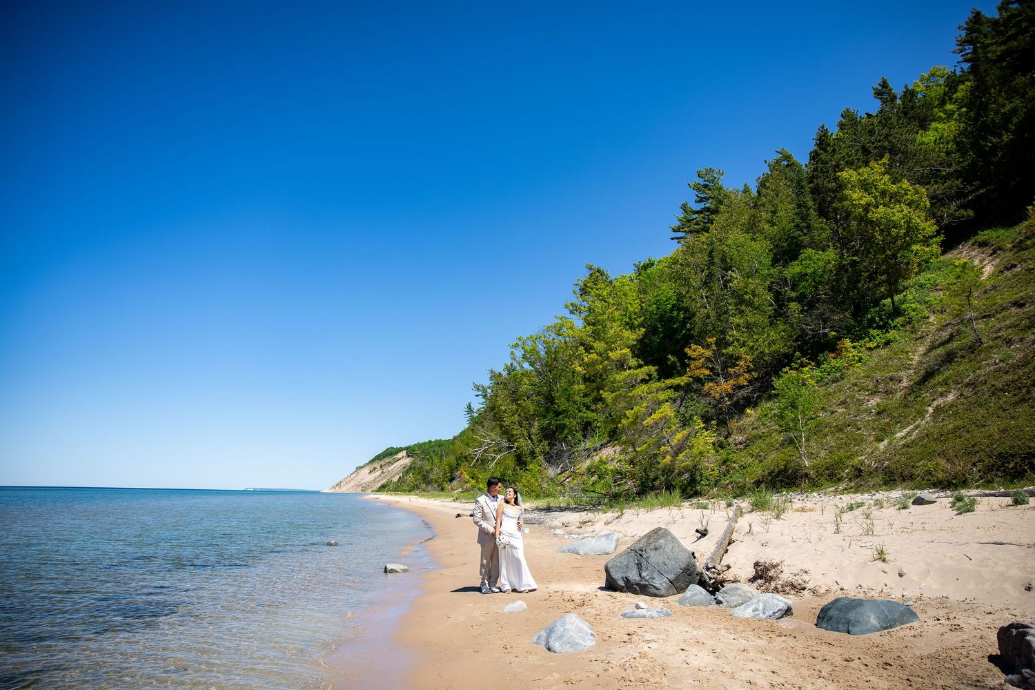 Couple in white attire standing on a sandy beach with clear blue water, green trees, and rocky cliffs under a bright blue sky.