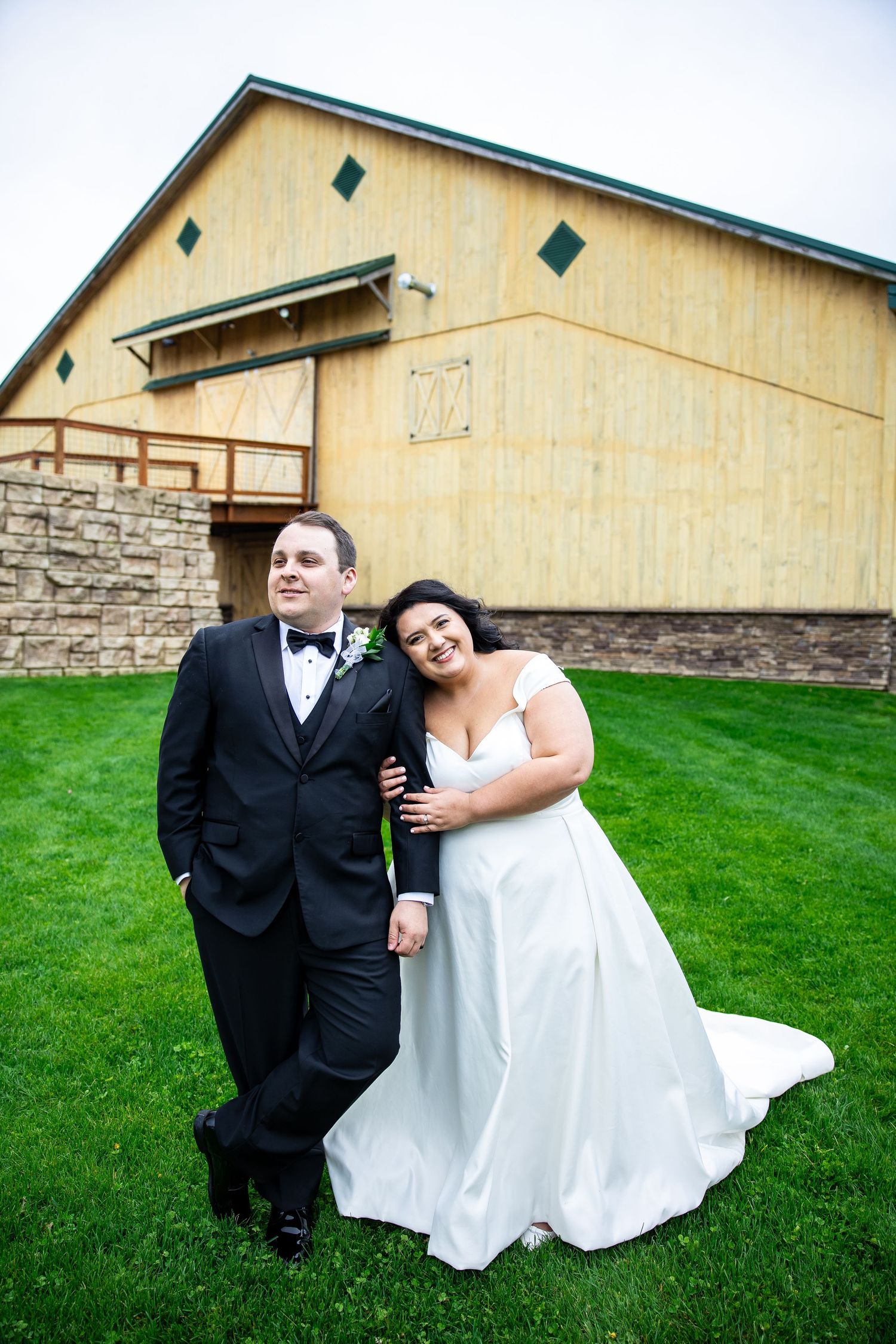 A bride in a white gown and groom in a black tuxedo pose together on green grass in front of a rustic barn venue.