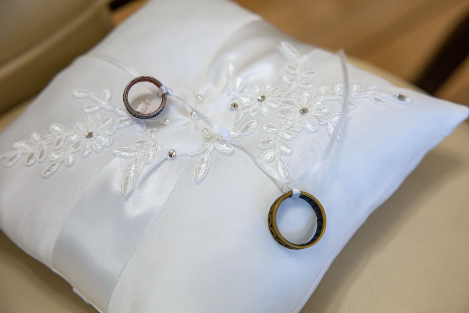 Two gold wedding rings resting on a white satin ring pillow with delicate lace embroidery and ribbon.