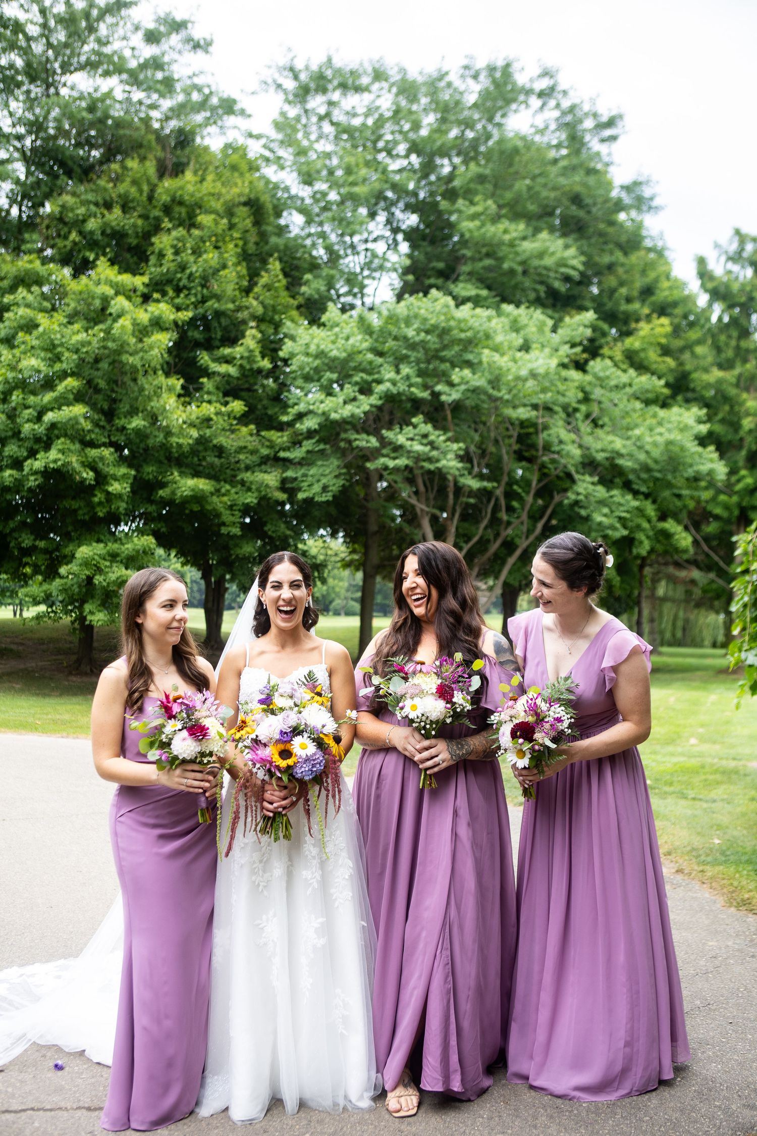 Bride in white gown laughs with three bridesmaids in purple dresses holding colorful bouquets outdoors.