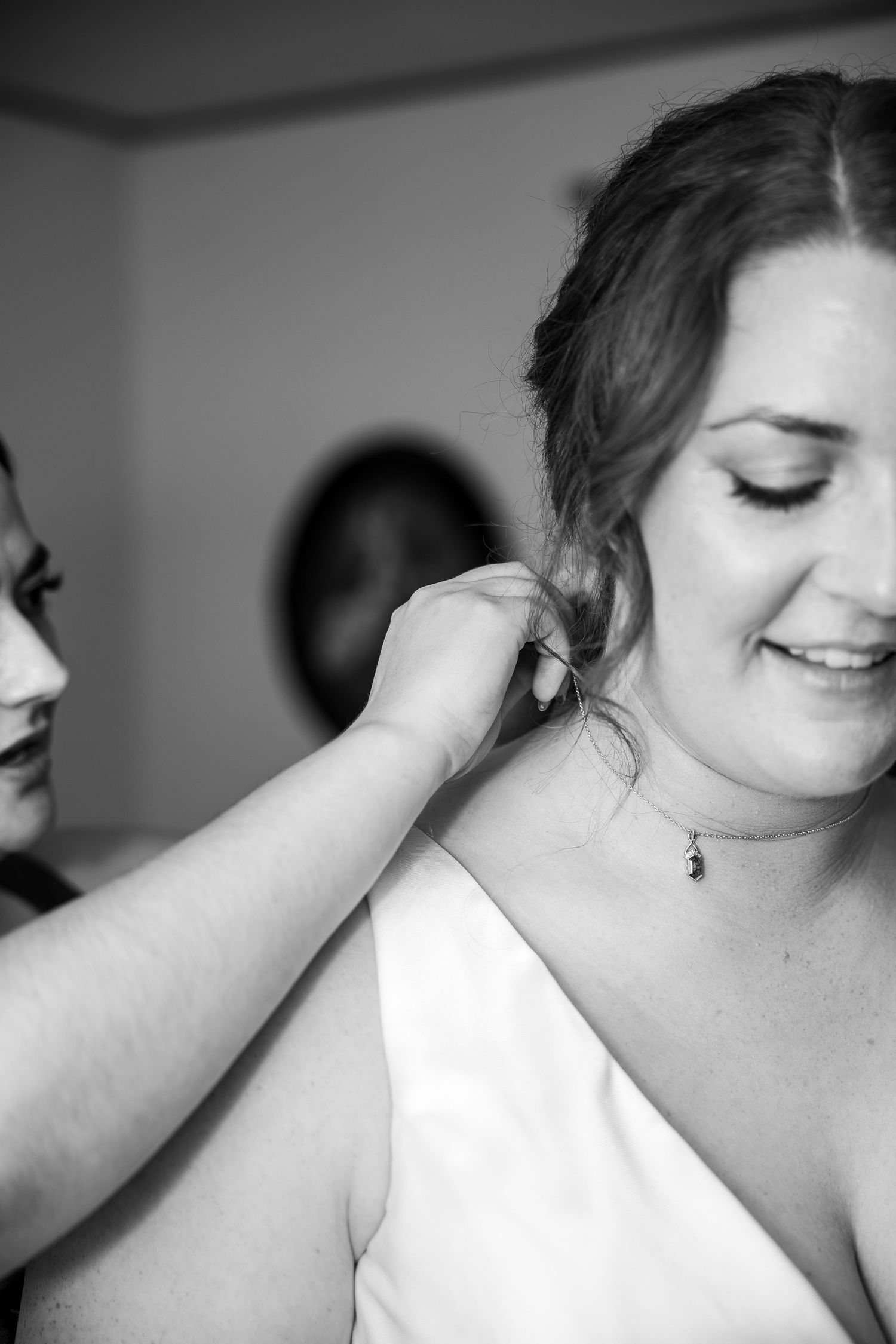 Black and white photo of a smiling bride in a white dress as a bridesmaid helps fasten her pendant necklace before the wedding.