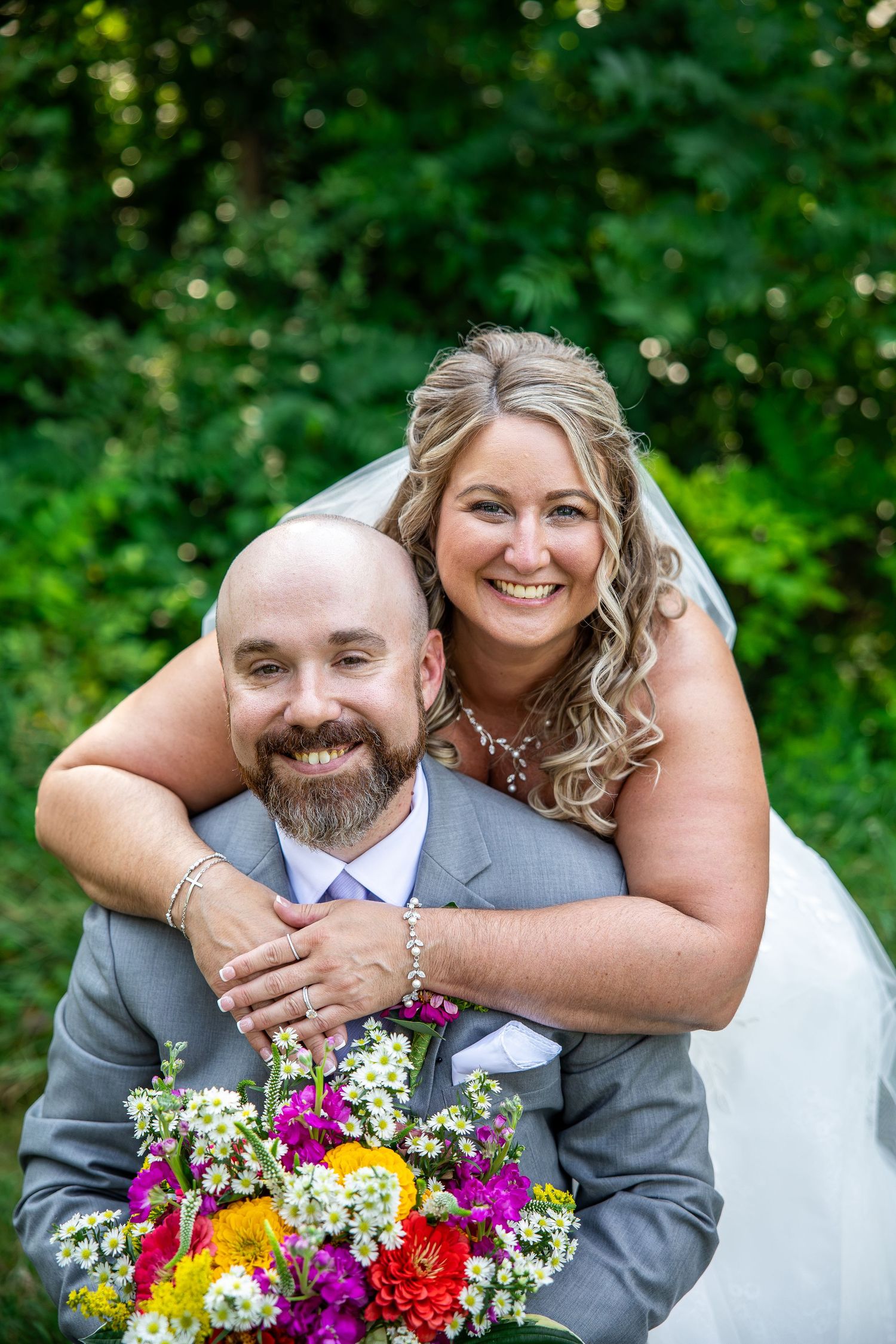Smiling bride in a white veil hugs her bearded groom in a gray suit from behind as he holds a colorful bouquet outdoors.