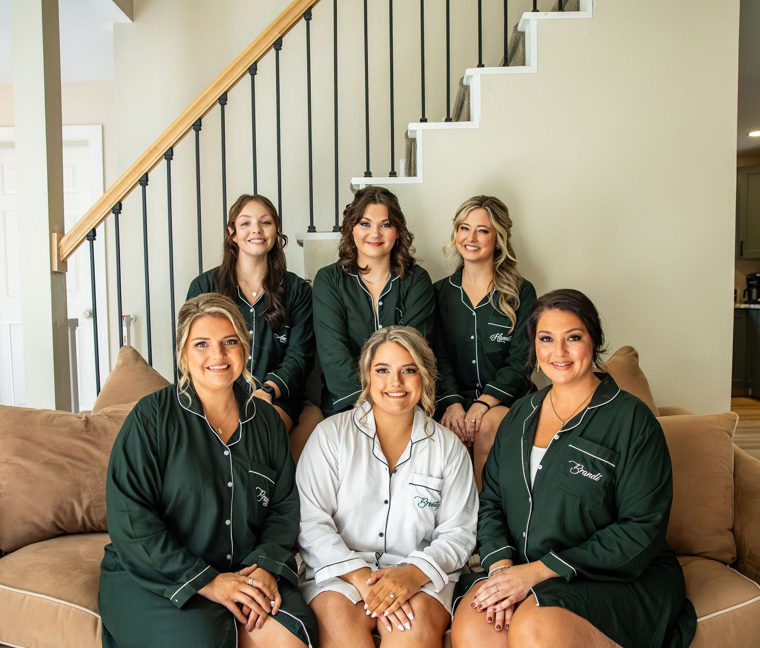 Six women in matching green and white button-up pajamas pose together on a tan sofa in front of a staircase, getting ready for a wedding.