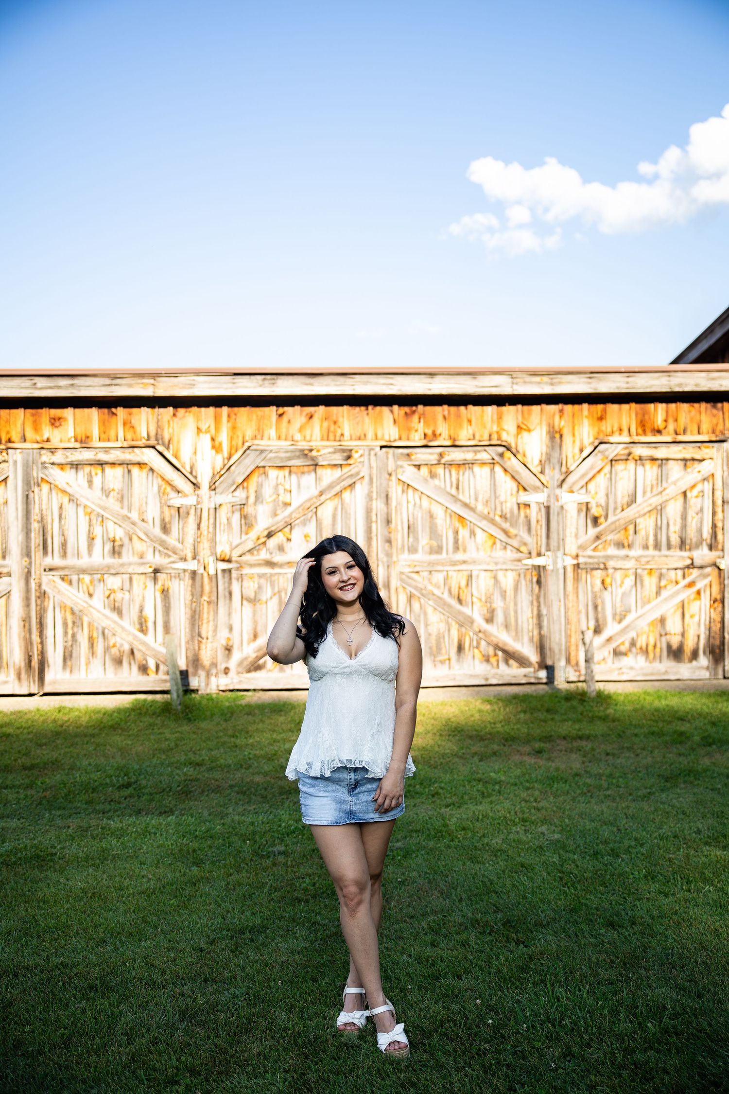 Young woman in white top and denim shorts smiling in front of rustic wooden barn doors on green grass.