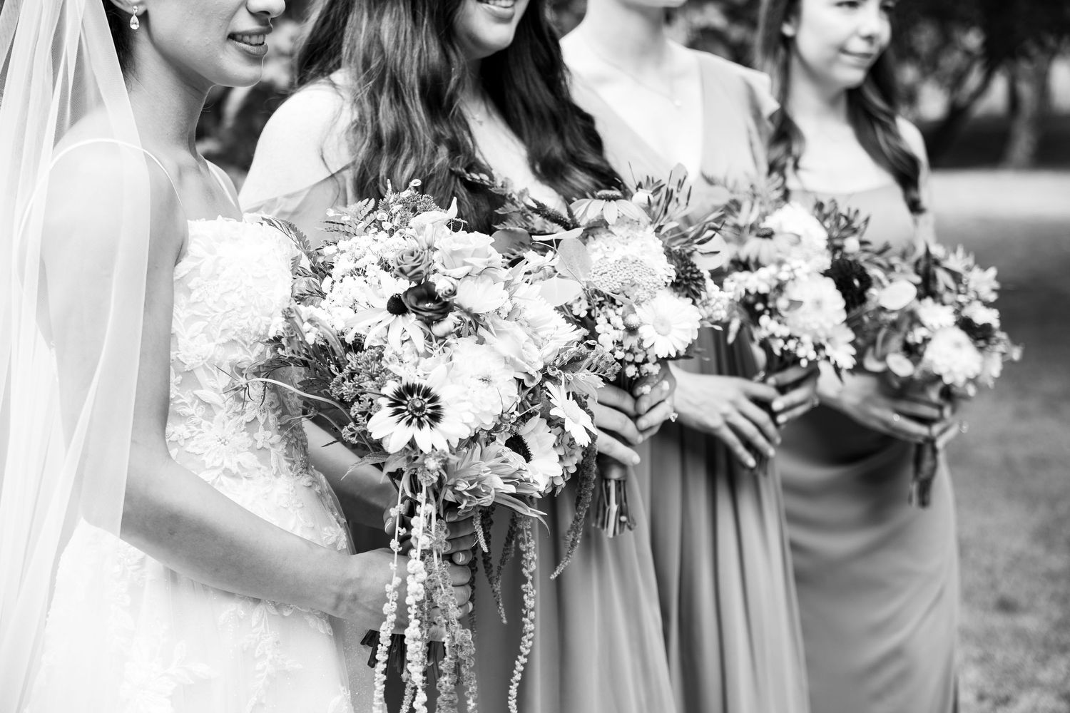 Black and white photo of a bride and bridesmaids holding lush floral bouquets at an outdoor wedding ceremony.