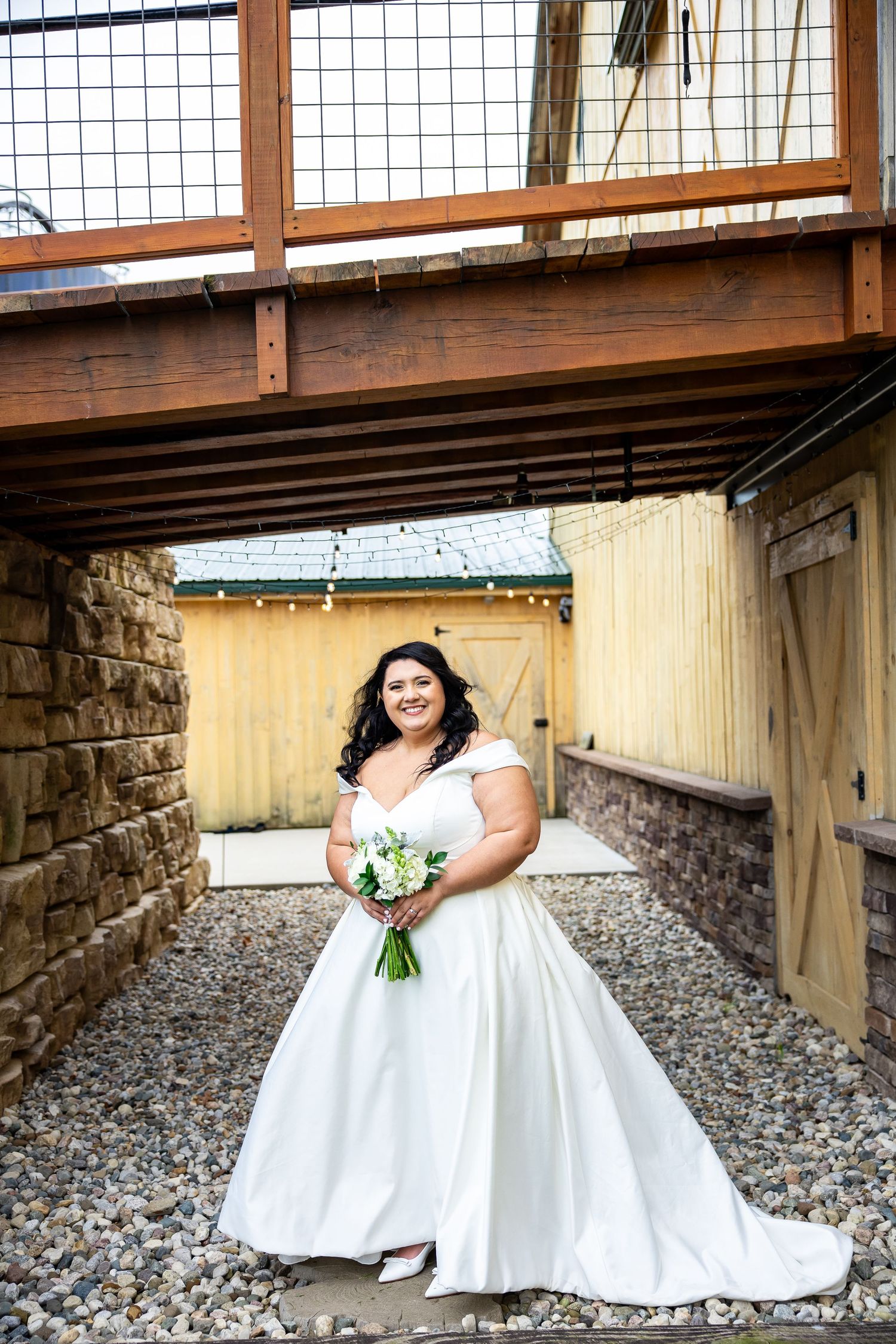 Smiling bride in white off-shoulder gown holding bouquet, posing on gravel path beneath wooden structure at rustic venue.