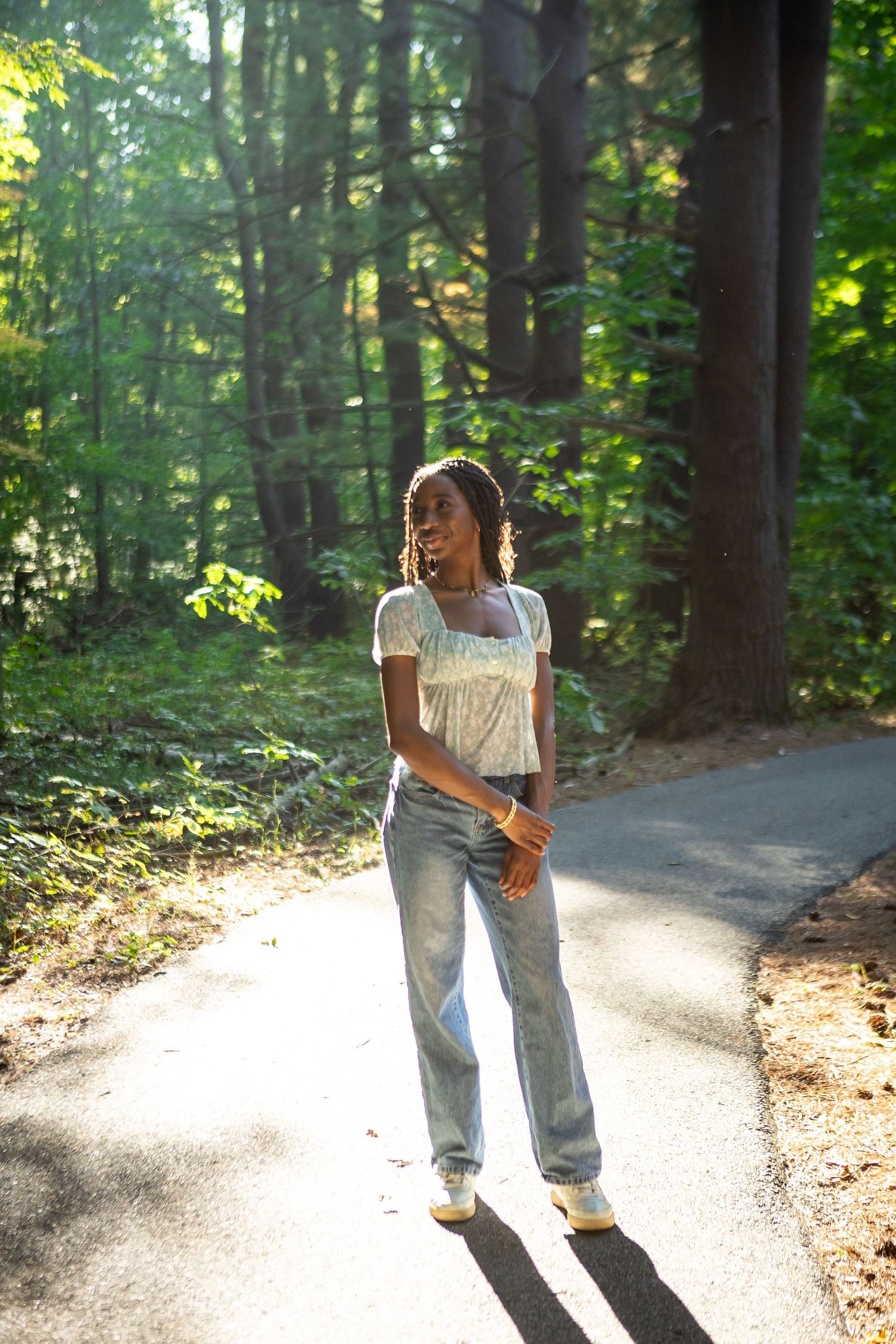 Person in casual white top and jeans stands on sunlit forest path during golden hour.