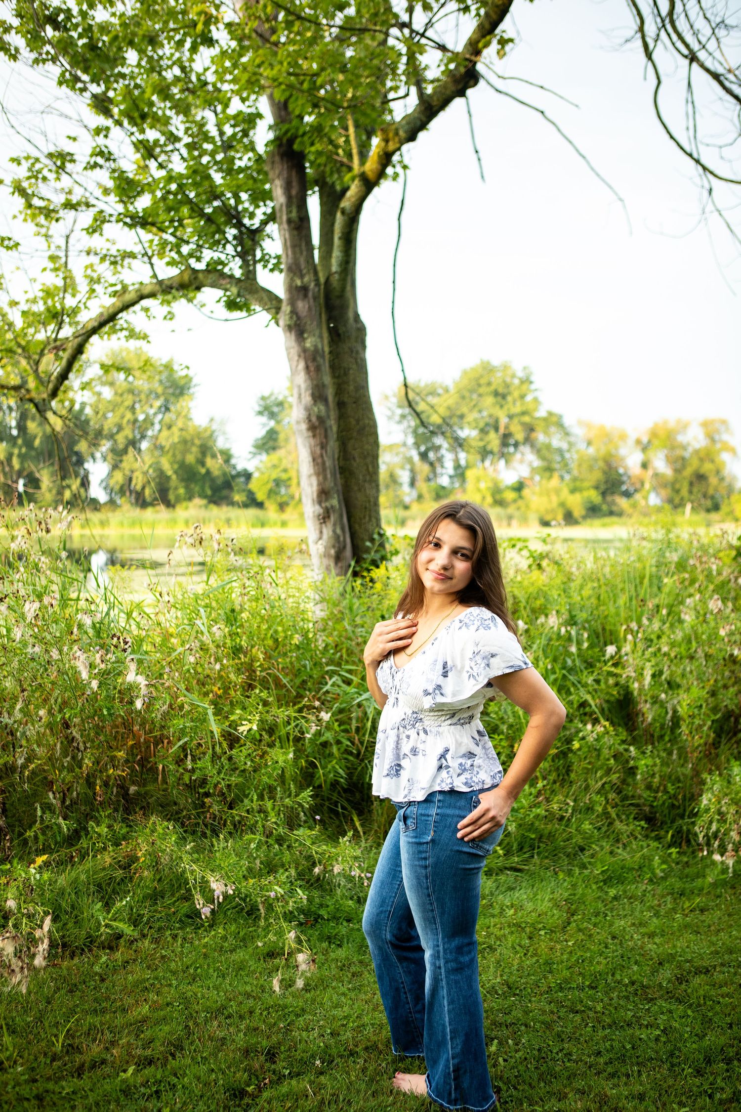 Young woman in floral top and jeans poses outdoors near a large tree with lush green meadow background.