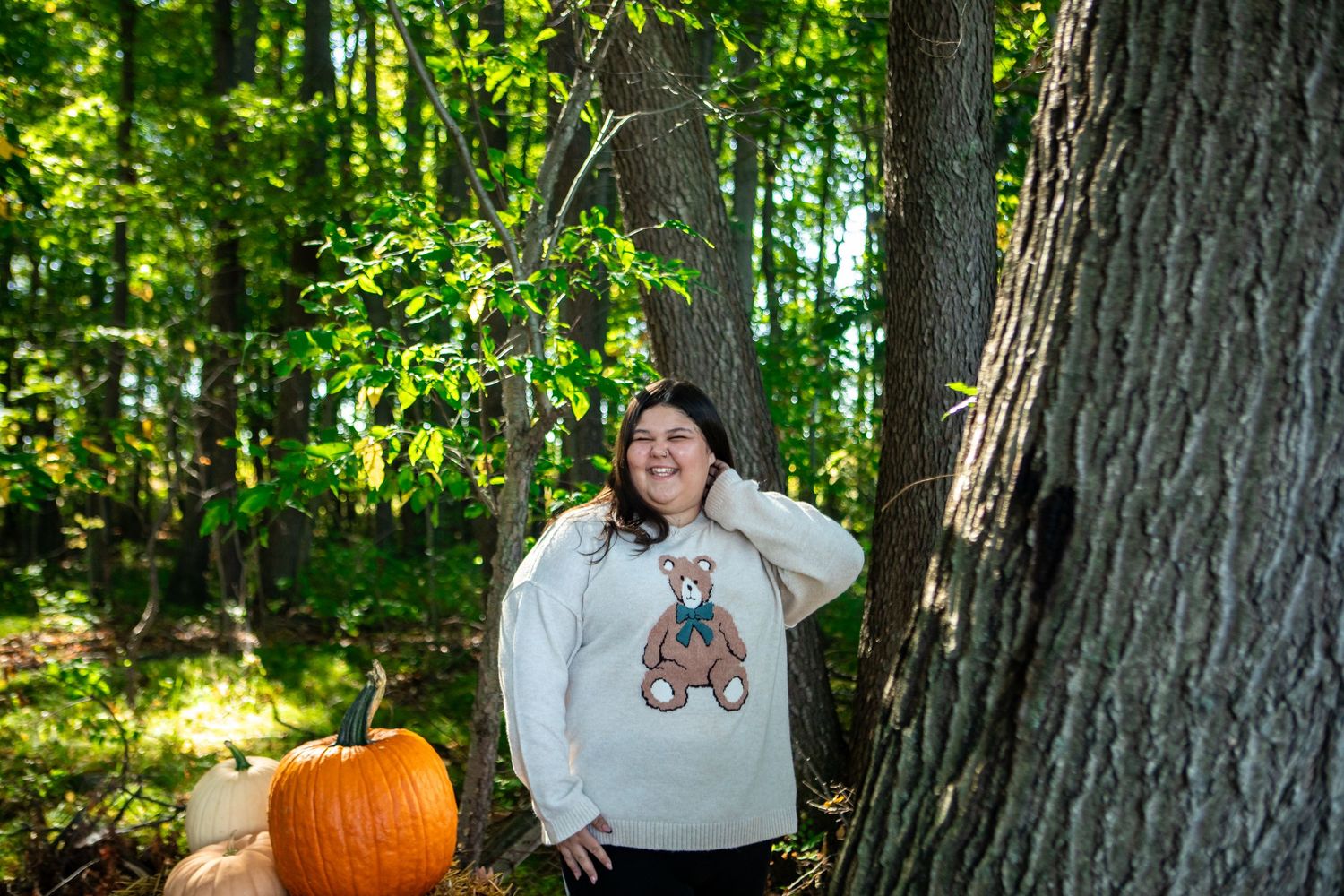 Woman in bear graphic sweatshirt poses by trees with orange pumpkin in autumn forest setting.