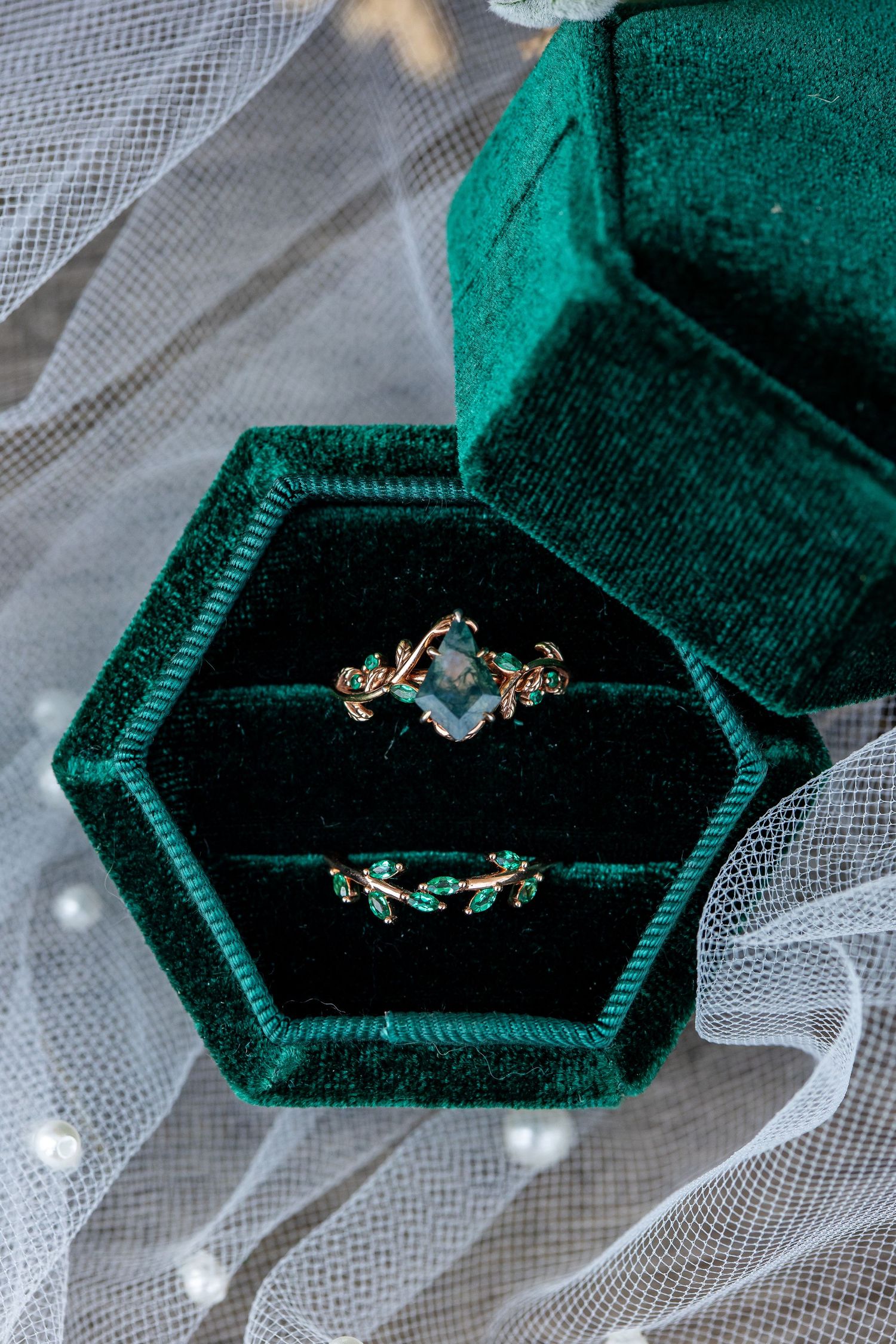 Green velvet hexagonal ring box holding a gold leaf-vine engagement ring with a moss agate stone and matching wedding band on white tulle.