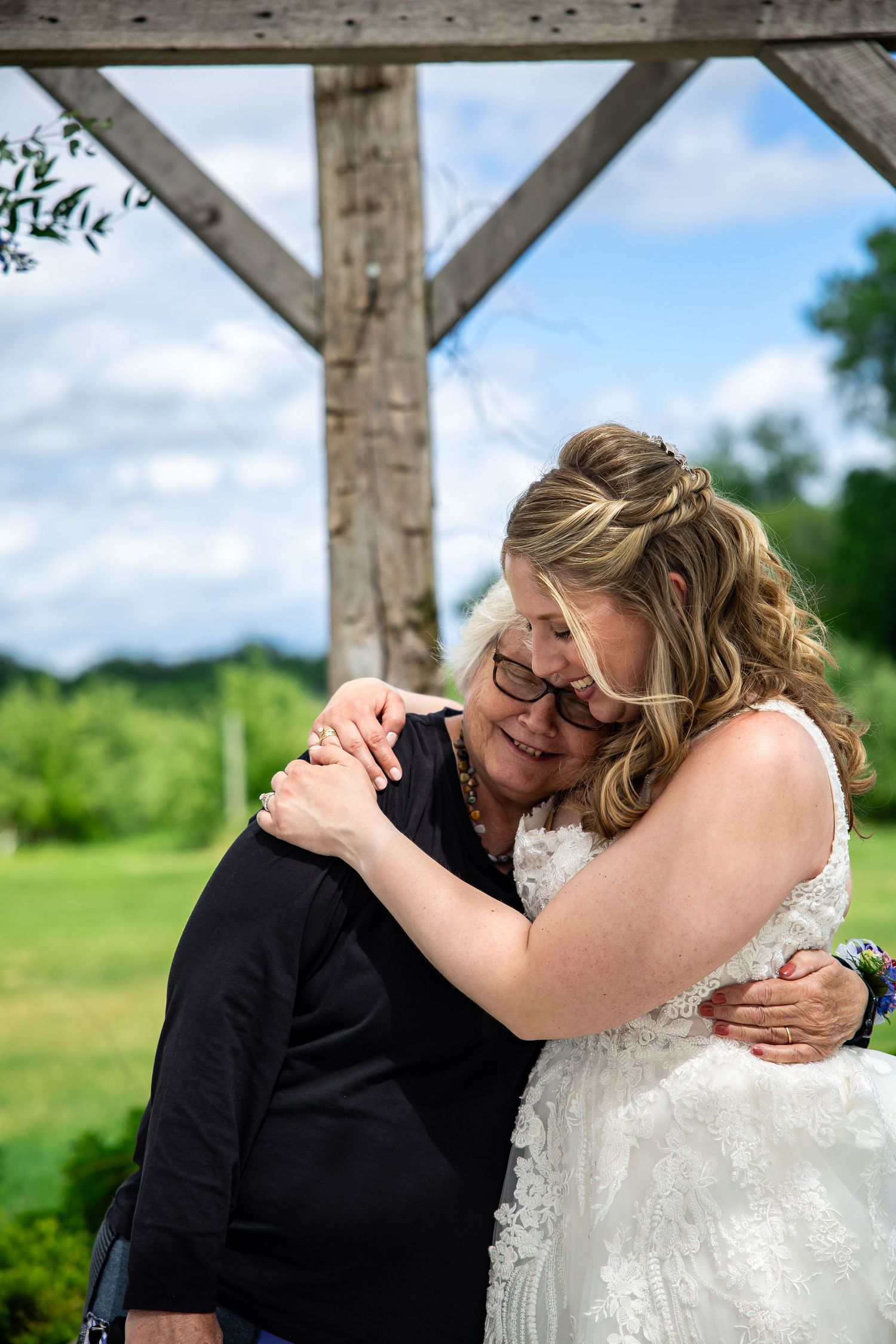 A bride in a white lace wedding dress shares a heartfelt embrace with an older woman in black under a rustic wooden pavilion.