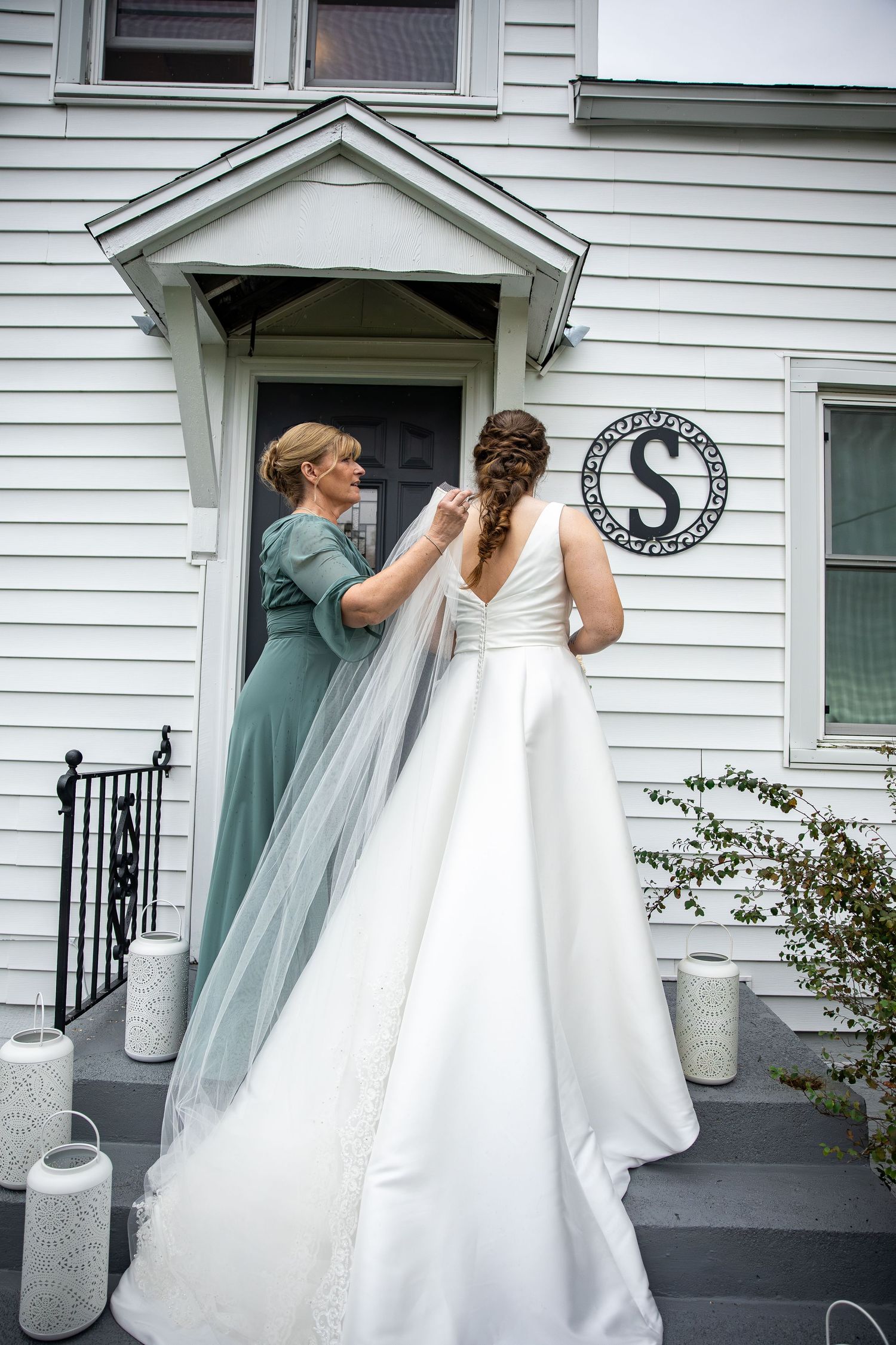 Woman in a green dress adjusts the veil and gown of a bride with a braided updo on the front steps of a white house.