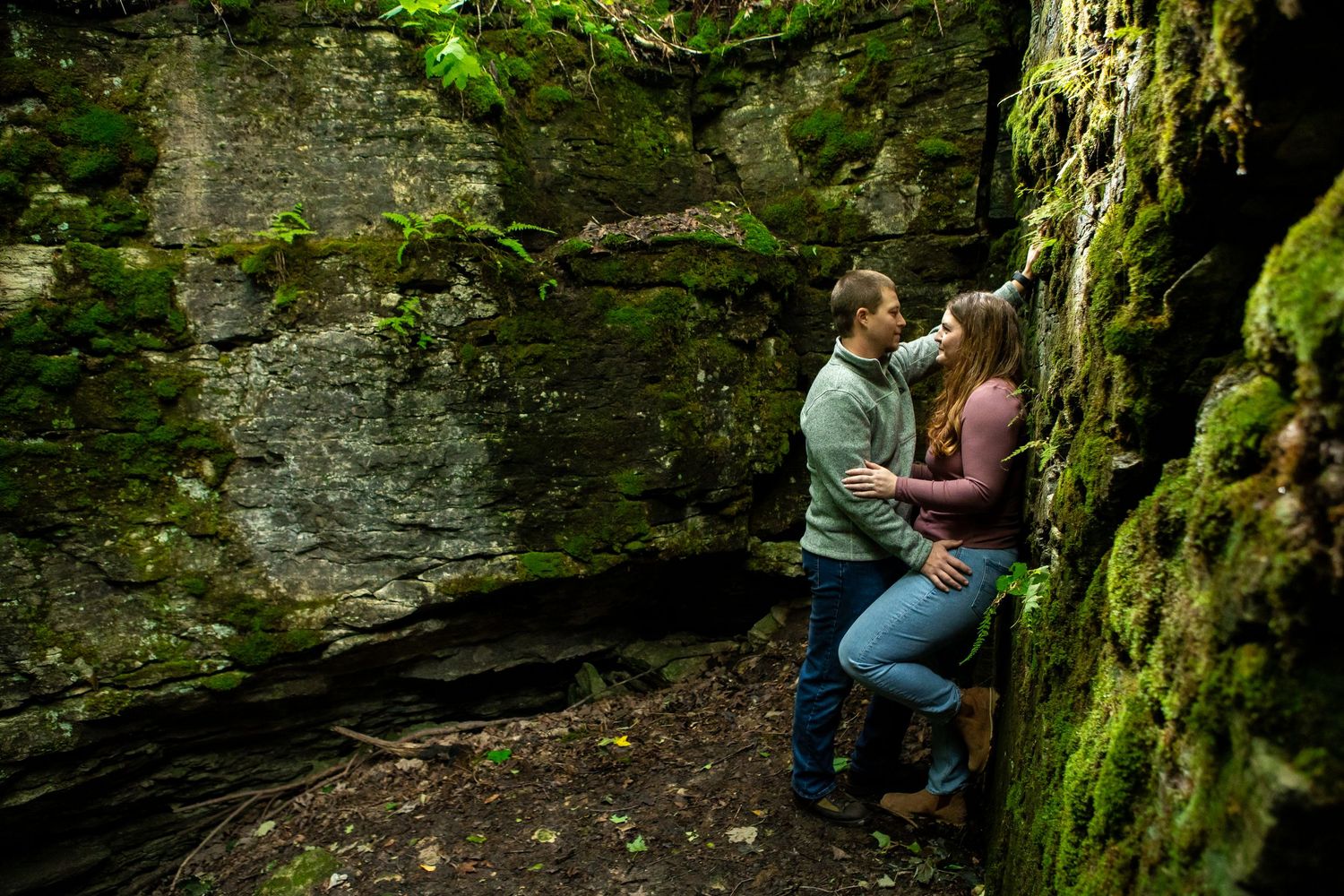 A couple embraces near a mossy rock wall in a natural setting.