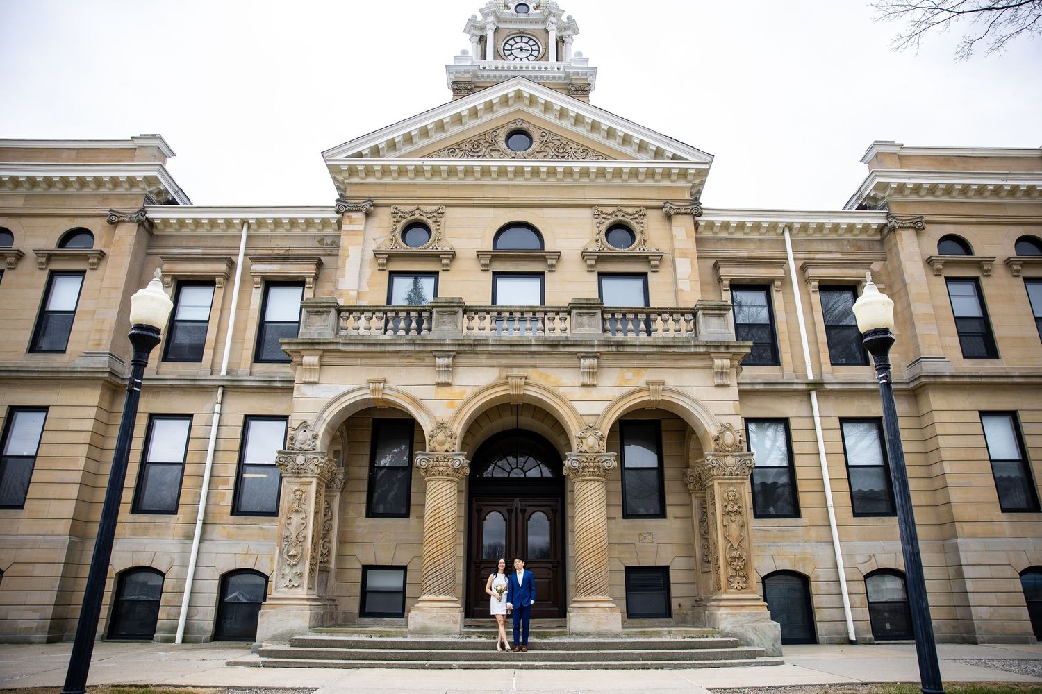 Couple poses on steps of historic limestone courthouse with ornate arched entrance and classical architecture.