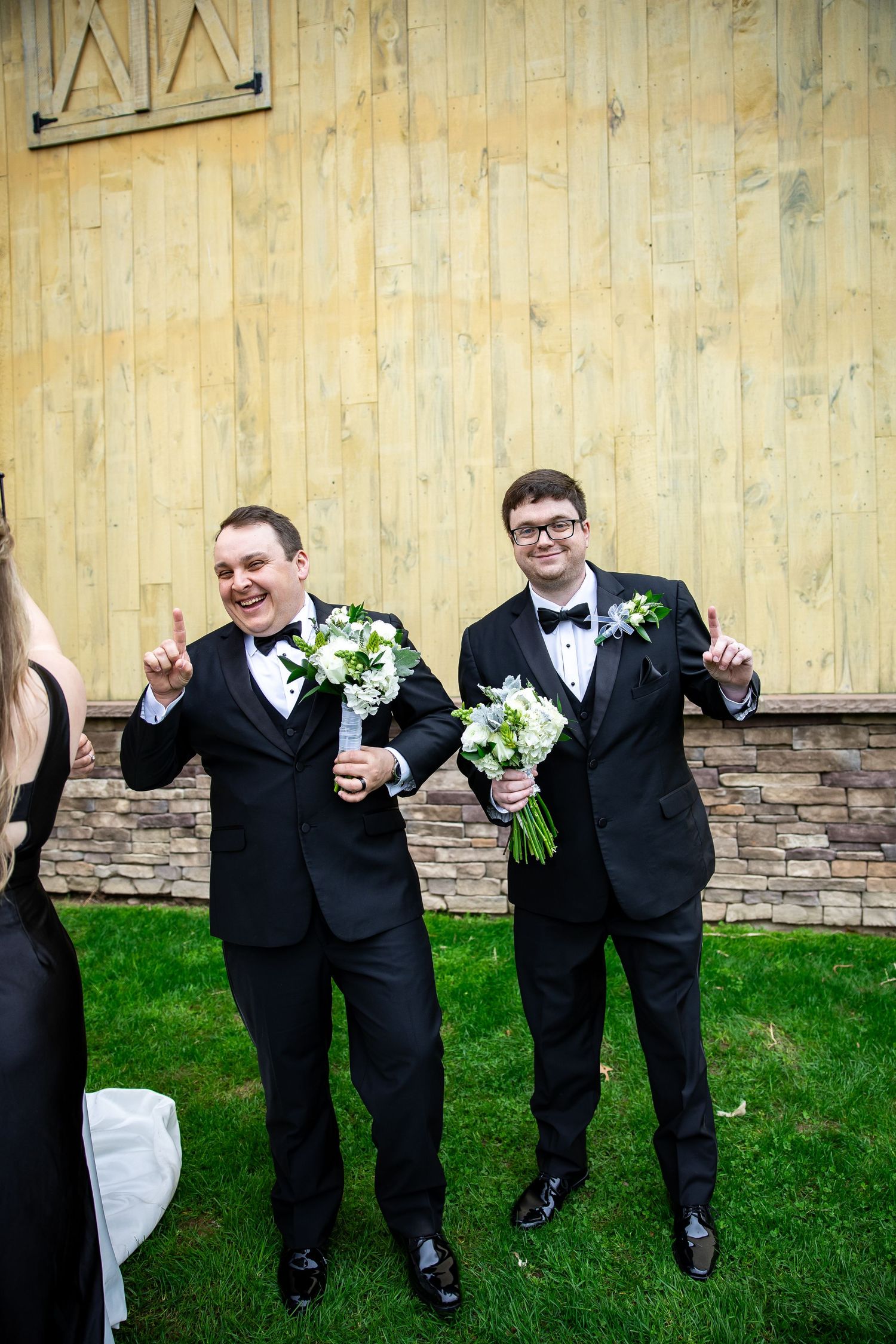Two groomsmen in black tuxedos holding white bouquets and pointing up, smiling outdoors near a wooden barn.