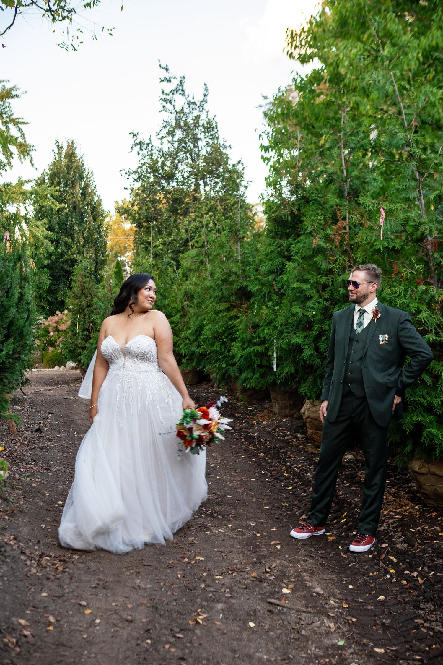 Bride in white gown and groom in dark suit share a joyful moment on a tree-lined path at an outdoor wedding.