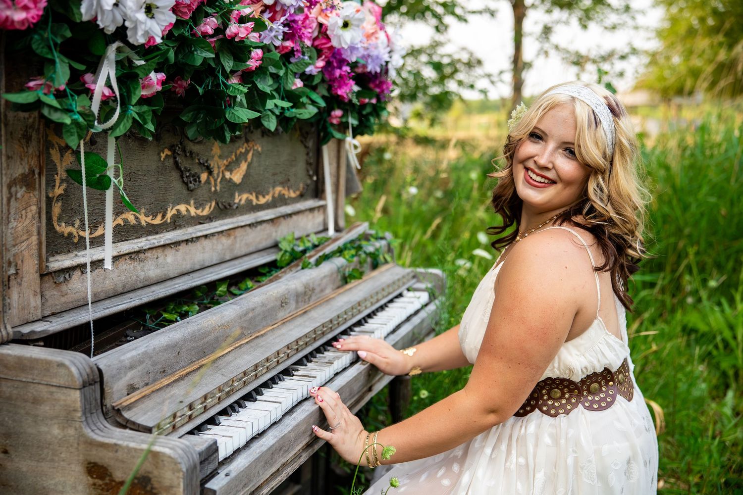 Smiling blonde woman in white dress playing a vintage upright piano outdoors surrounded by pink and white flowers.