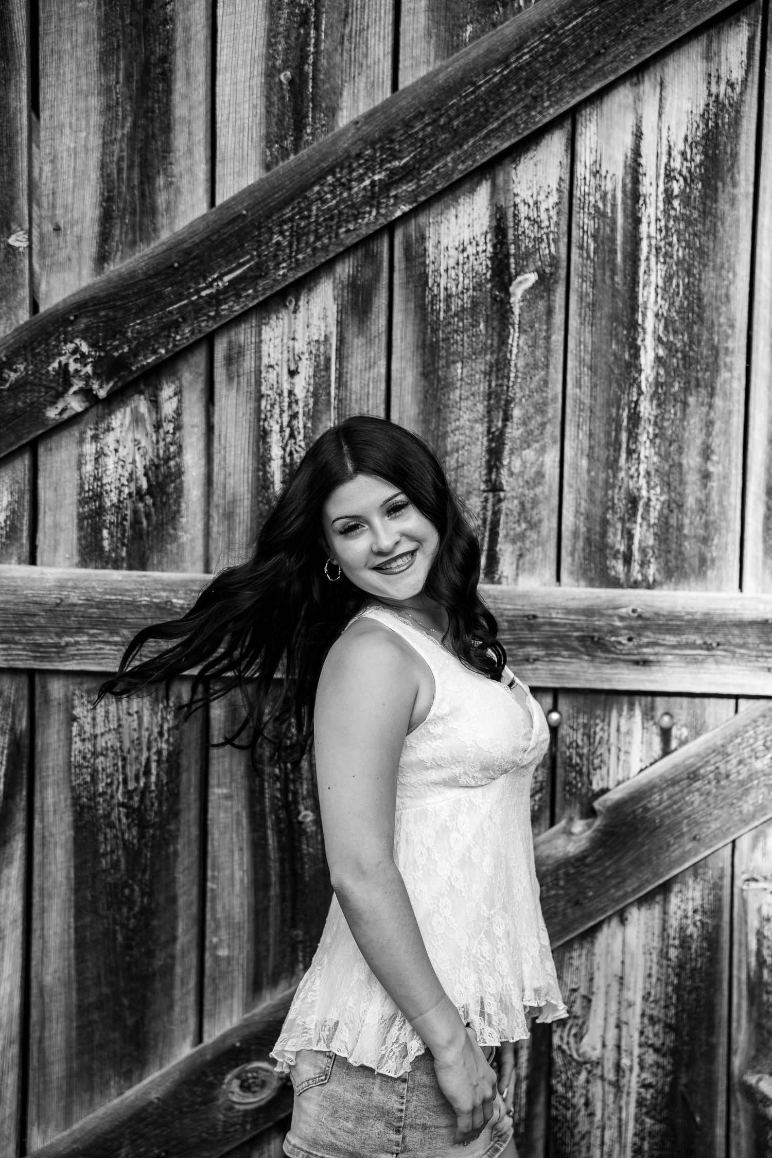 Black and white portrait of a smiling young woman with flowing dark hair leaning against a rustic wooden barn wall.