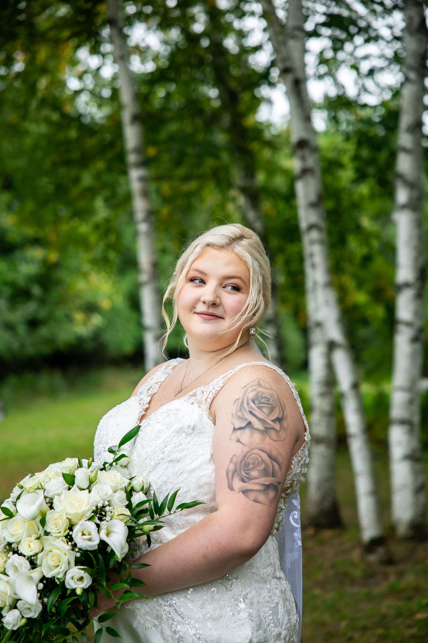 Smiling bride in a white lace wedding gown with rose tattoos holds a white floral bouquet among birch trees outdoors.