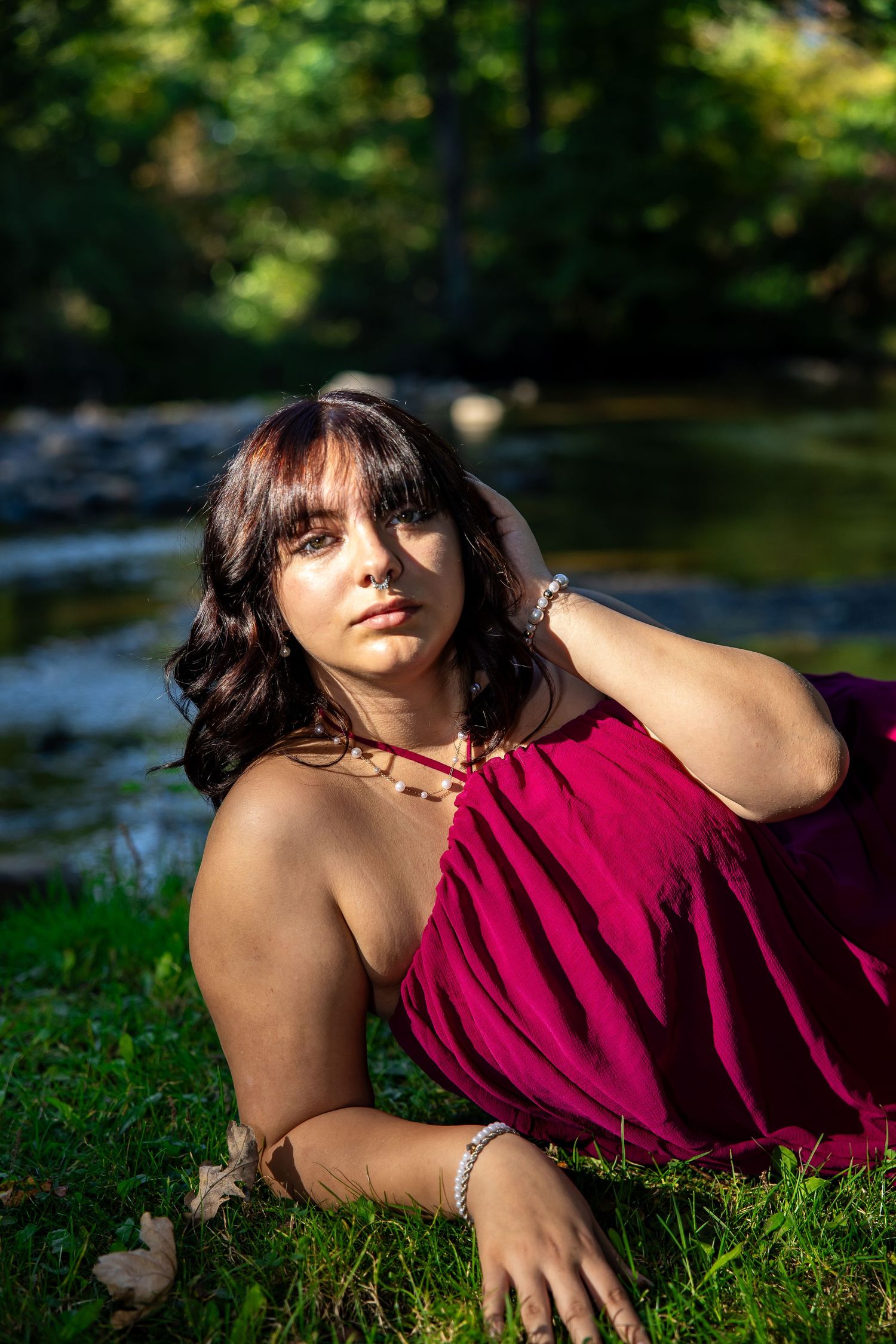 Young woman in magenta dress reclining on grass by a stream, gazing upward in a sunlit outdoor setting.