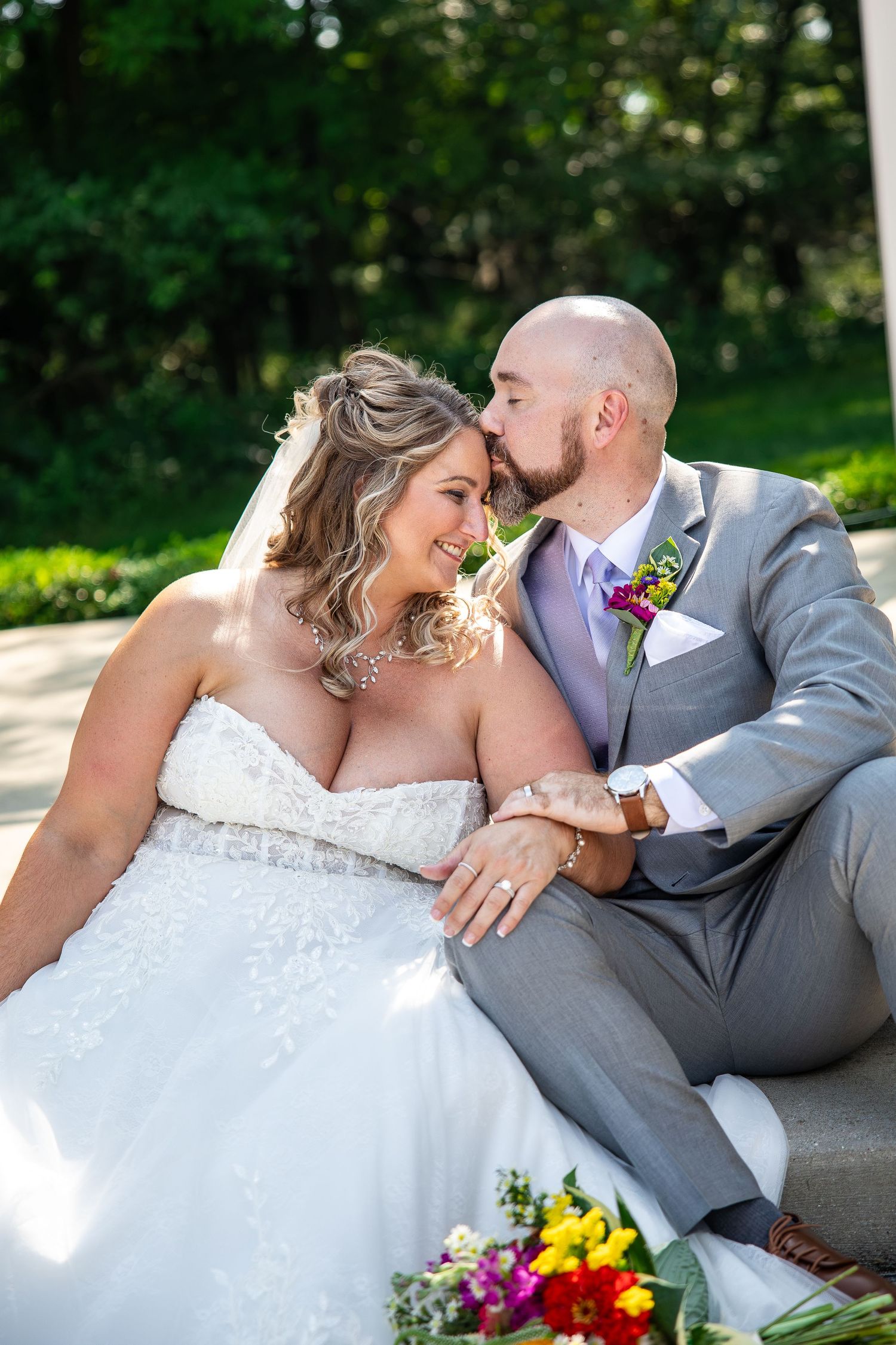 Groom in gray suit kisses bride in strapless lace gown on forehead while sitting outdoors with colorful bouquet.