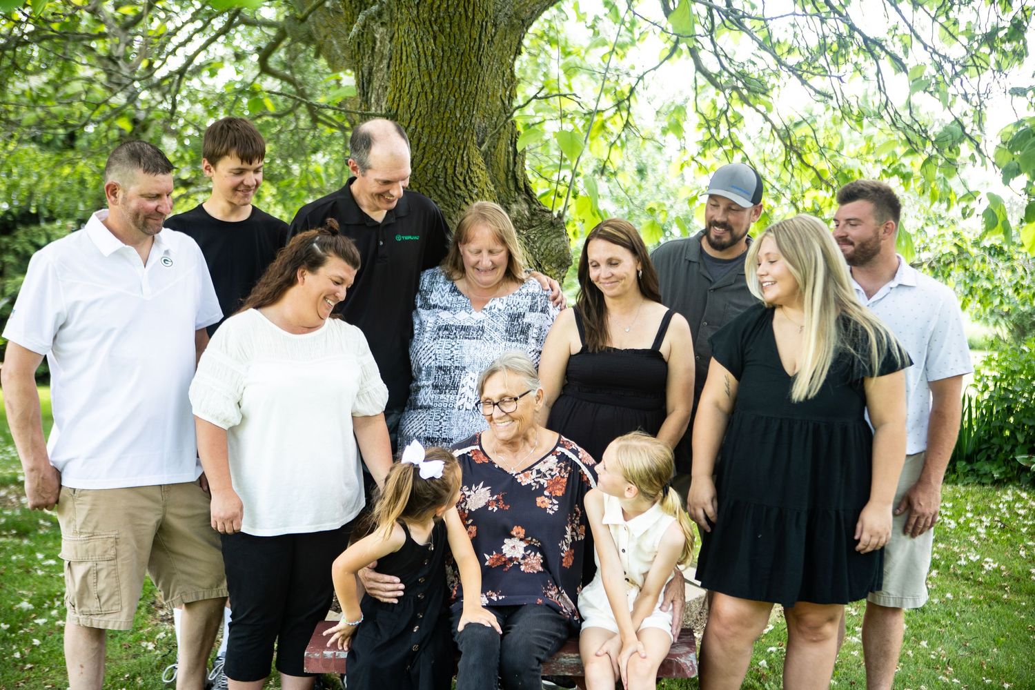 Family gathering under large tree branches in casual summer attire.