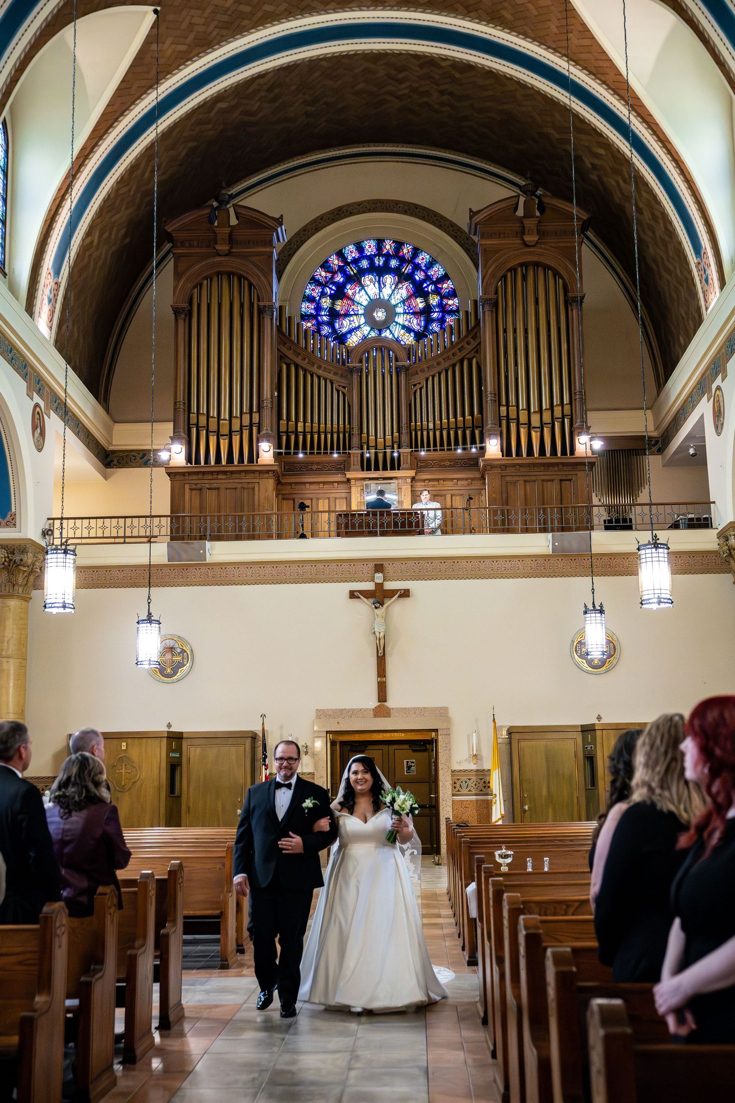 Bride walks down the aisle with escort in a beautiful church with stained glass rose window and pipe organ.