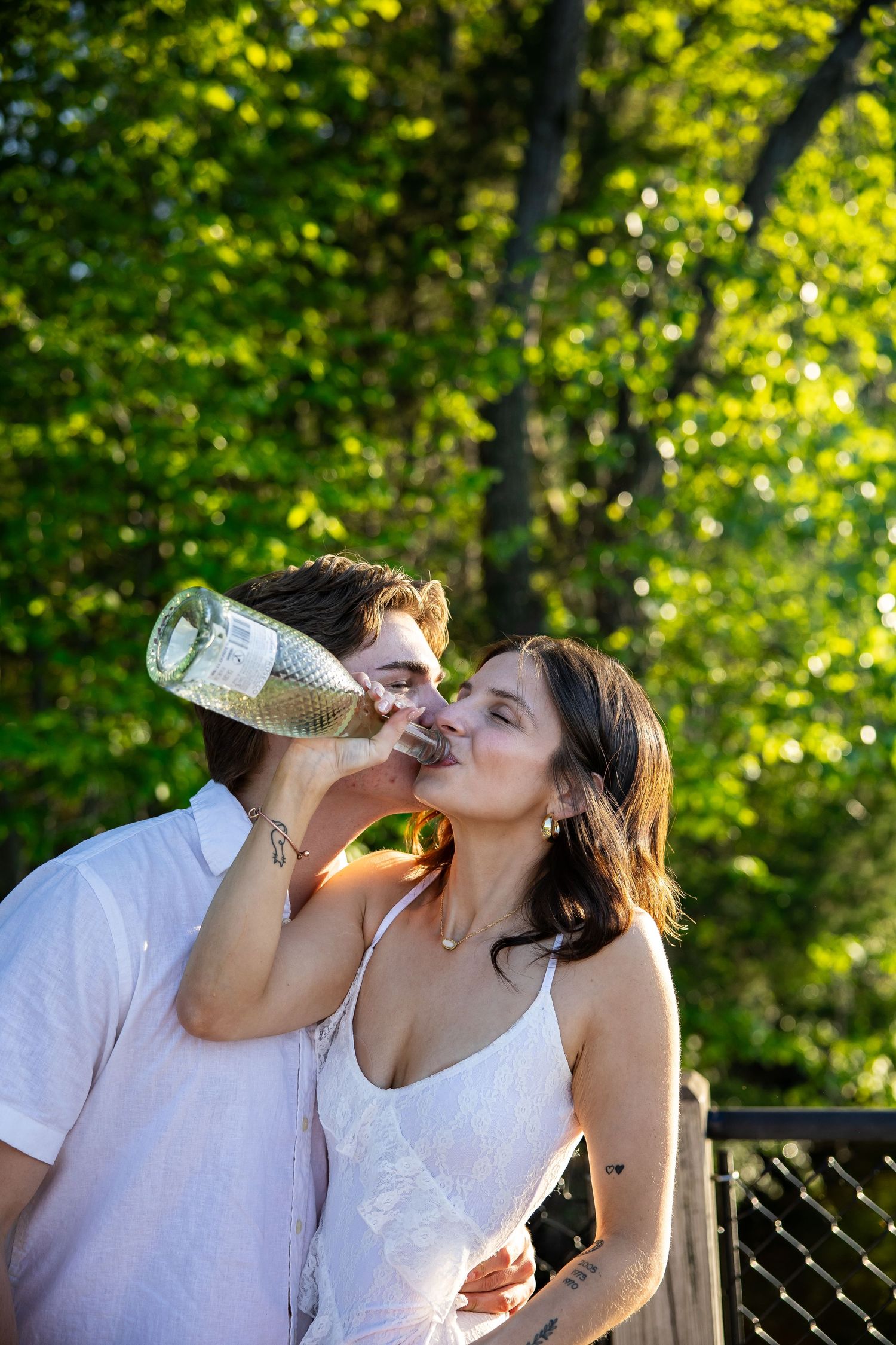 Couple sharing a drink from a wine bottle outdoors in a sunny green park setting.